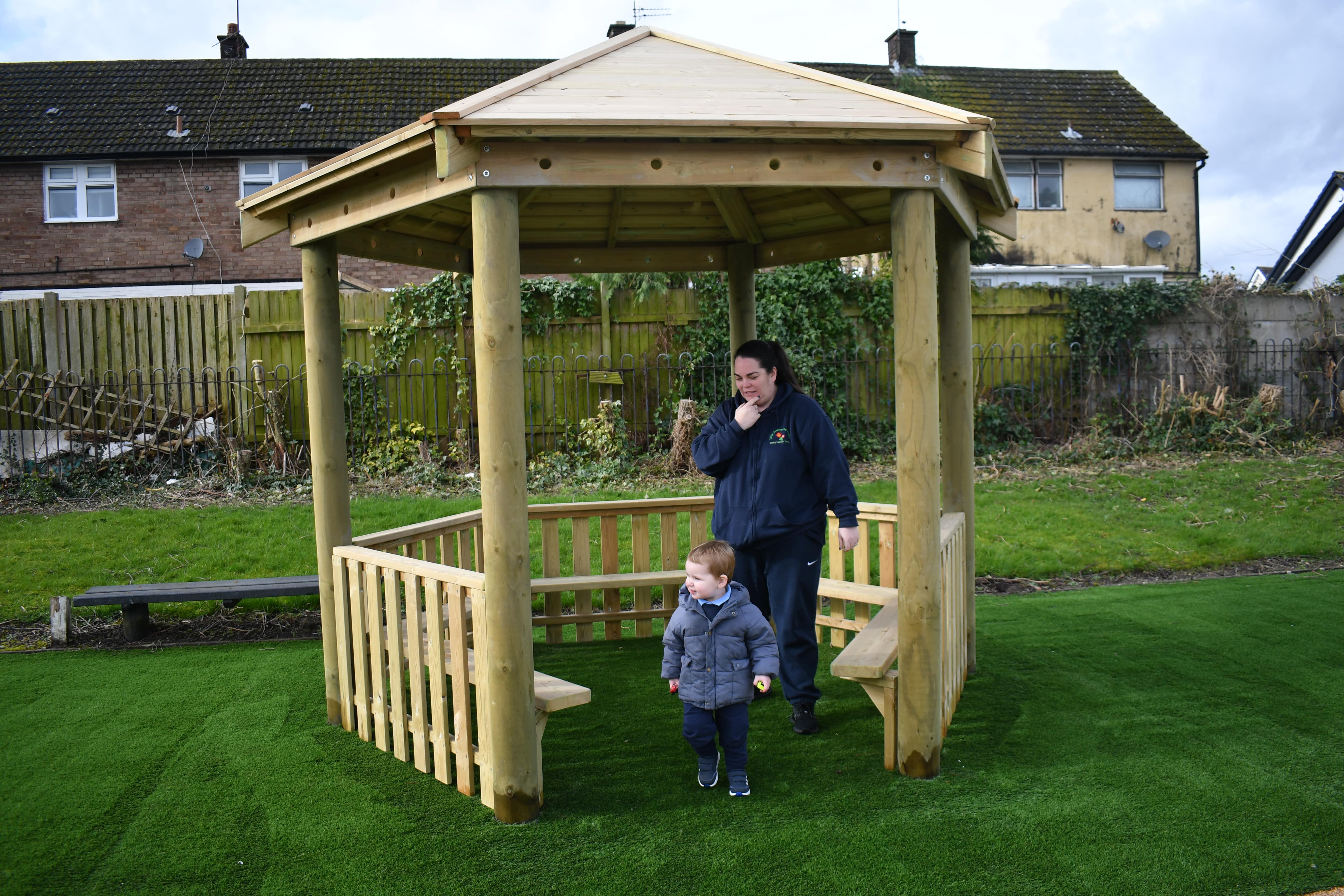 An adult is following a little child out of the Wooden Canopy. The canopy has been installed on artificial grass and houses can be seen in the background.