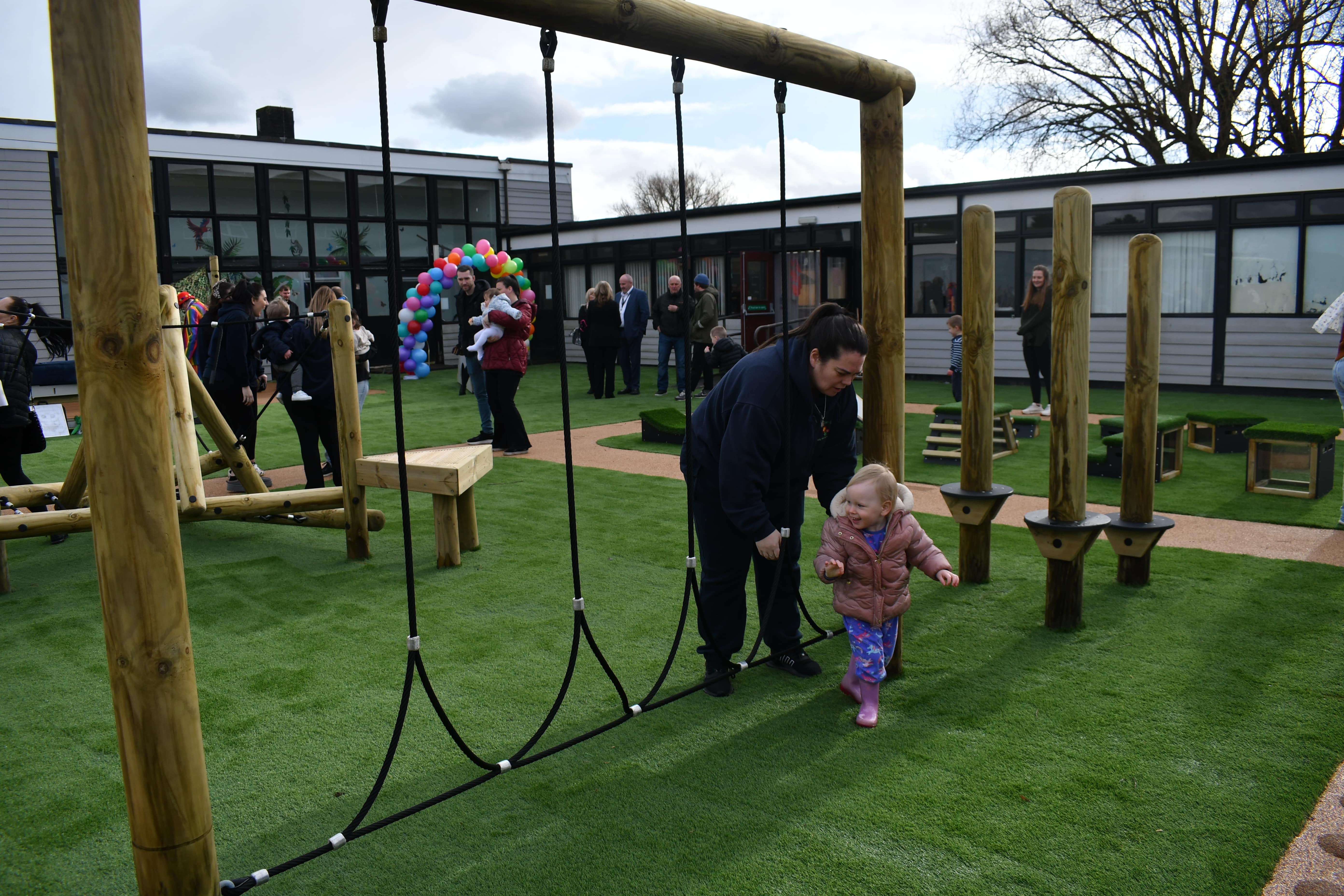 A little child is stood next to the Rope Log Traversal piece of equipment. An adult is following the child and helping them explore the new playground.