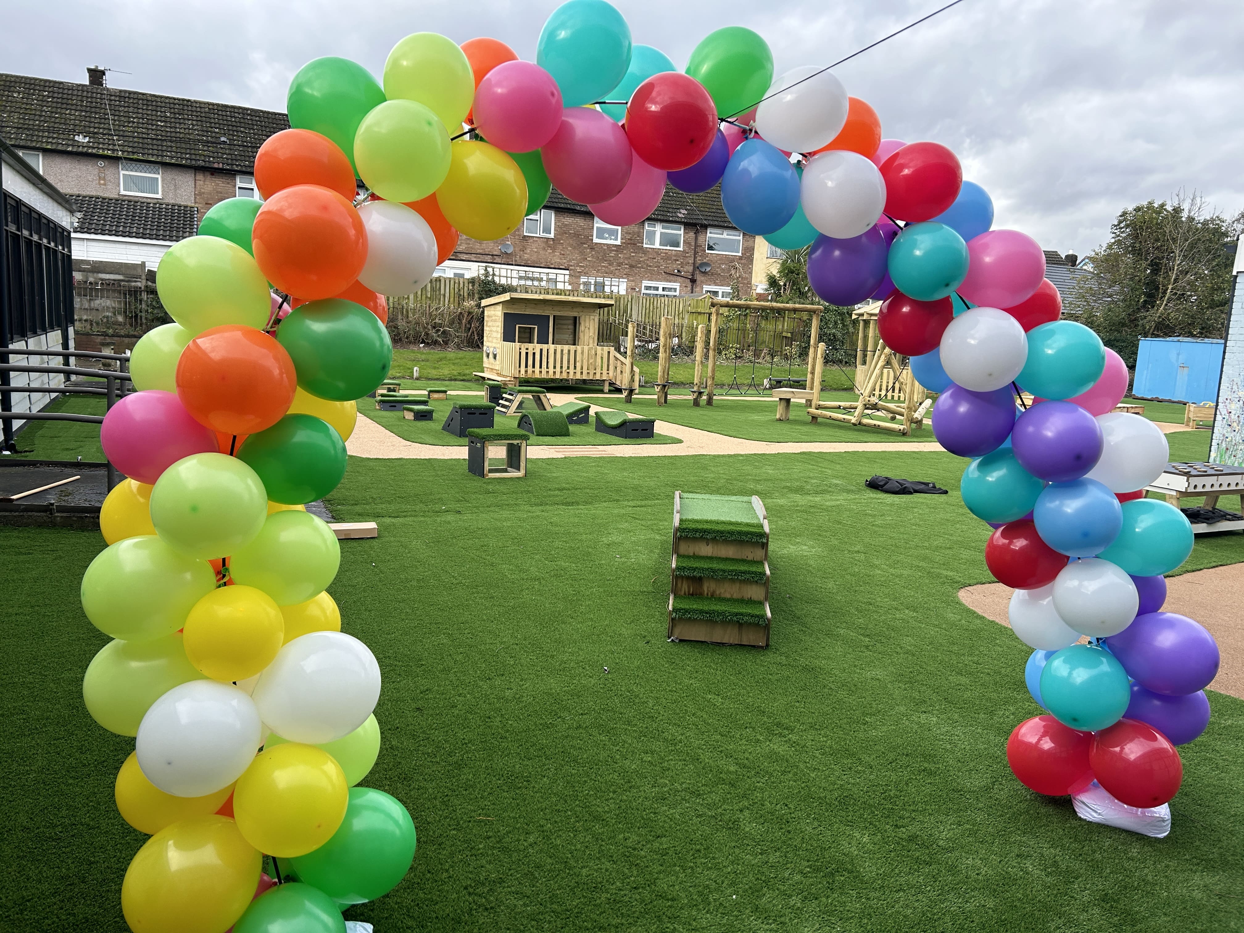 A balloon arch has been placed at the entrance to the play area. You can see a variety of commercial play equipment on the other side, with artificial grass and wetpour surfacing.