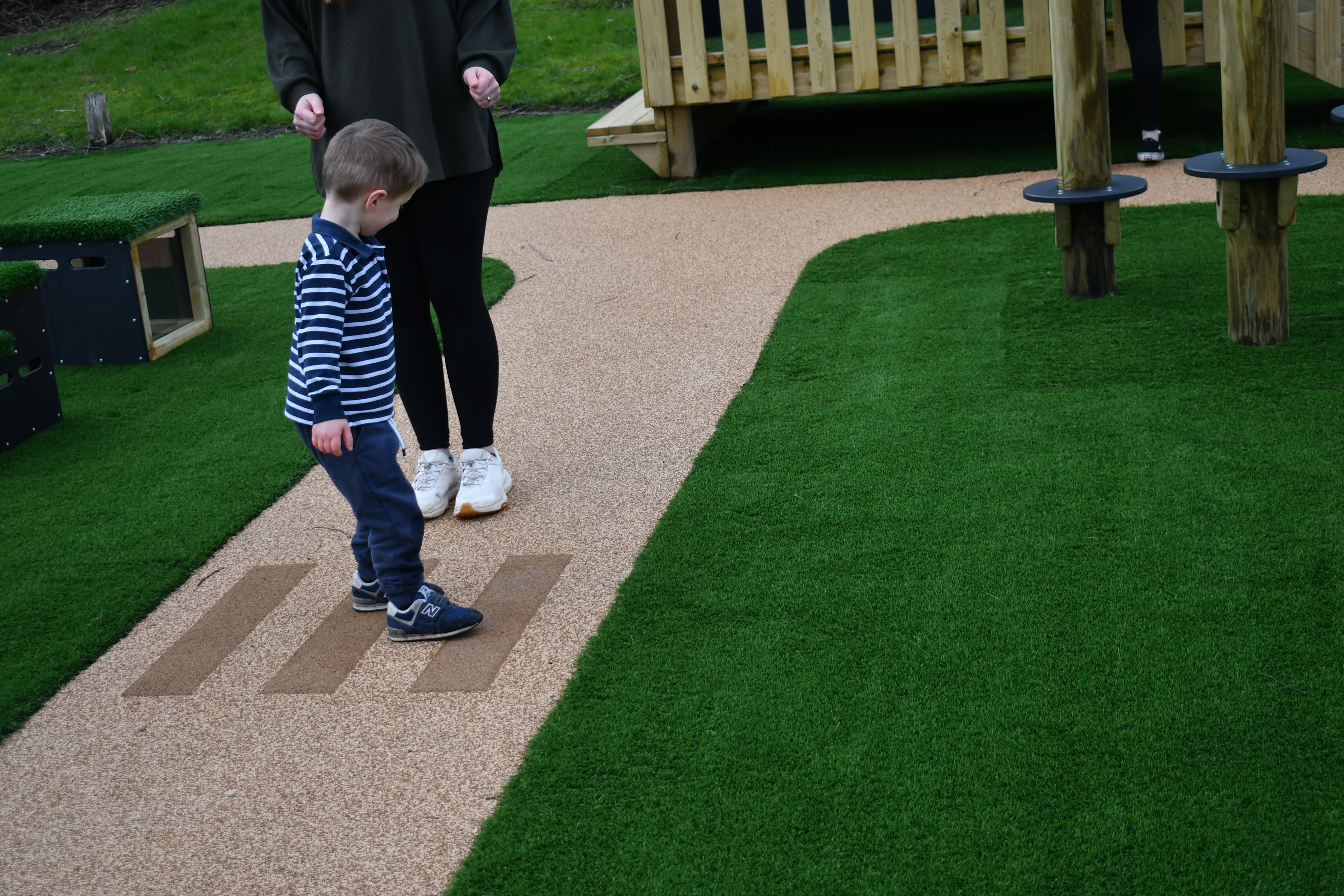 A small child is walking across a wetpour path, designed to look like a road. A zebra crossing marking has been installed on the wetpour surface. An adult is watching the child cross.