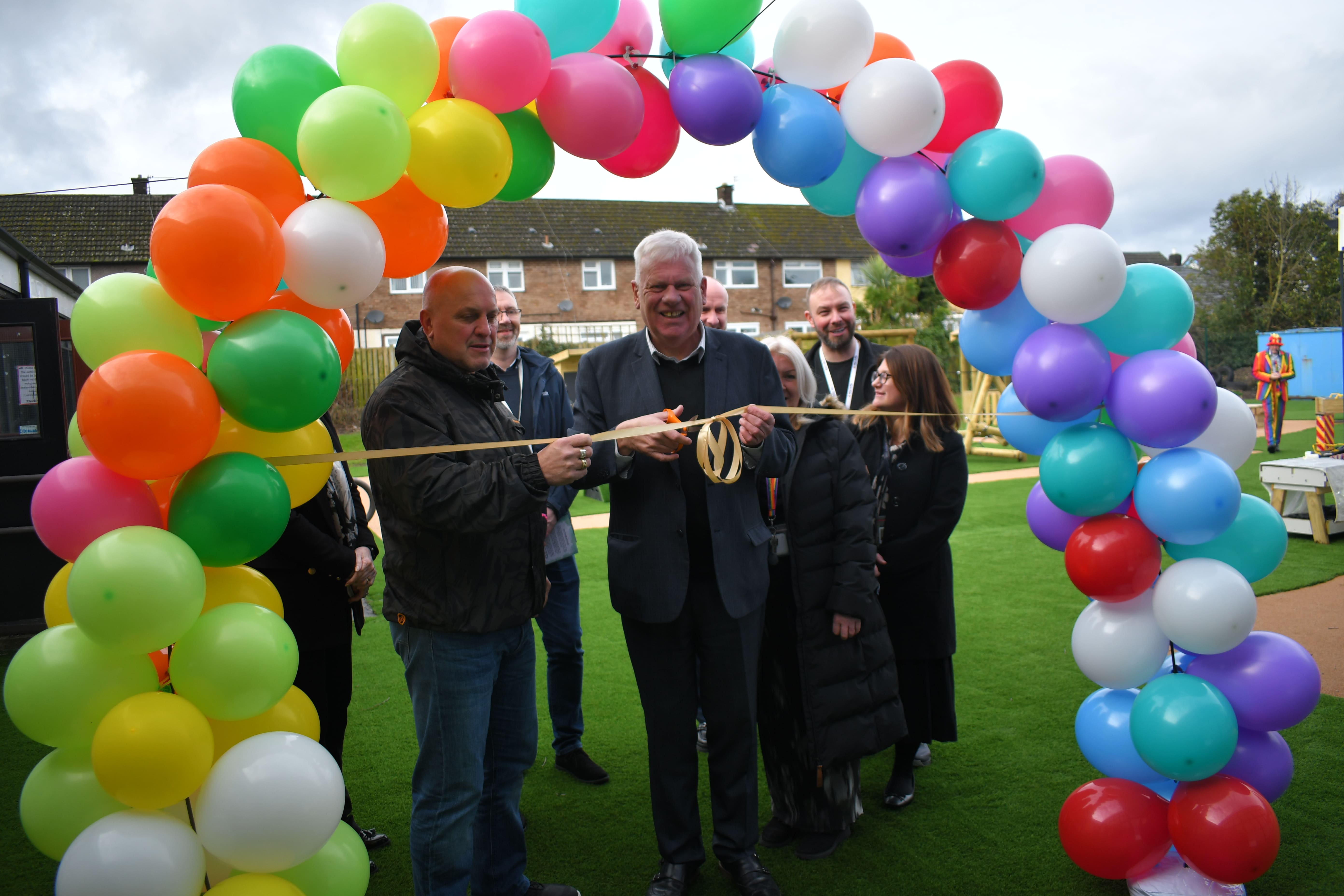 A group of adults are stood underneath a balloon arch, made up of multiple colours. A golden ribbon has been placed across the balloon arch as one adult holds a pair of scissors, ready to cut the ribbon.