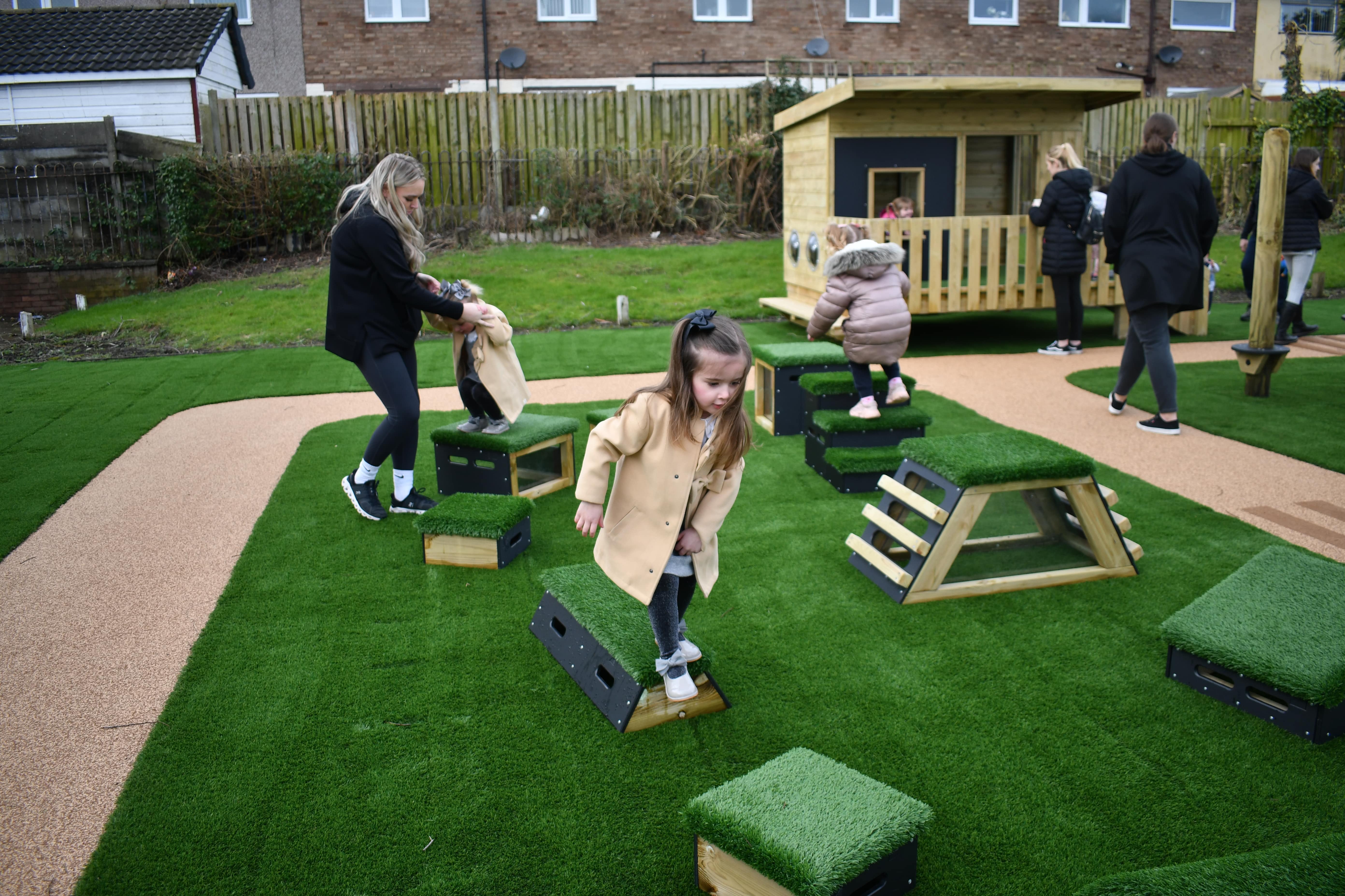 A group of children are playing on Get Set, Go! Blocks. These blocks are from the Mendips set and contain 12 different wooden blocks with artificial grass toppings. An adult is helping one child across them.