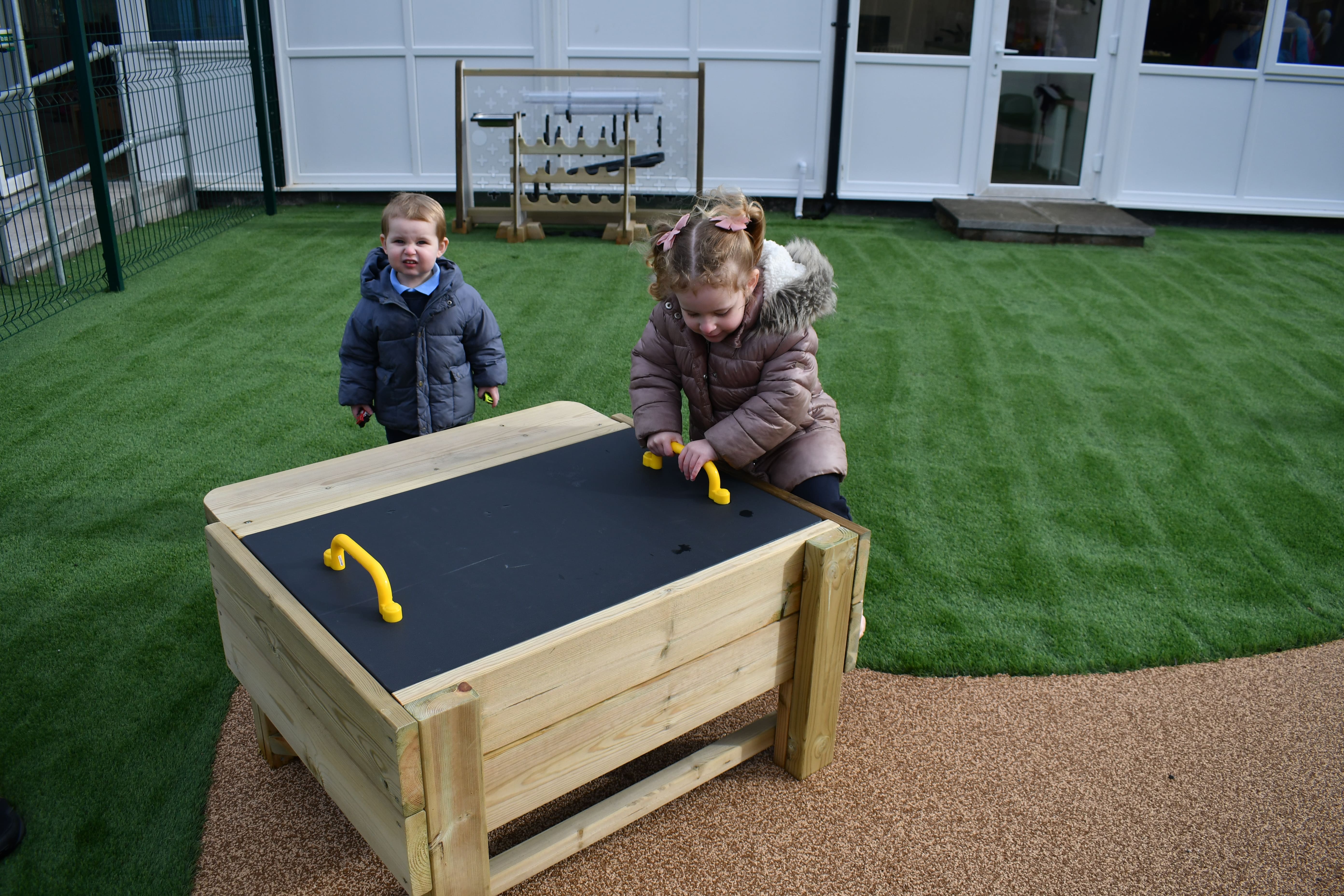 Two children are trying to take the lid off a box, which has been placed on a wetpour surface. One child is looking into the camera whilst the other tries to lift the lid.