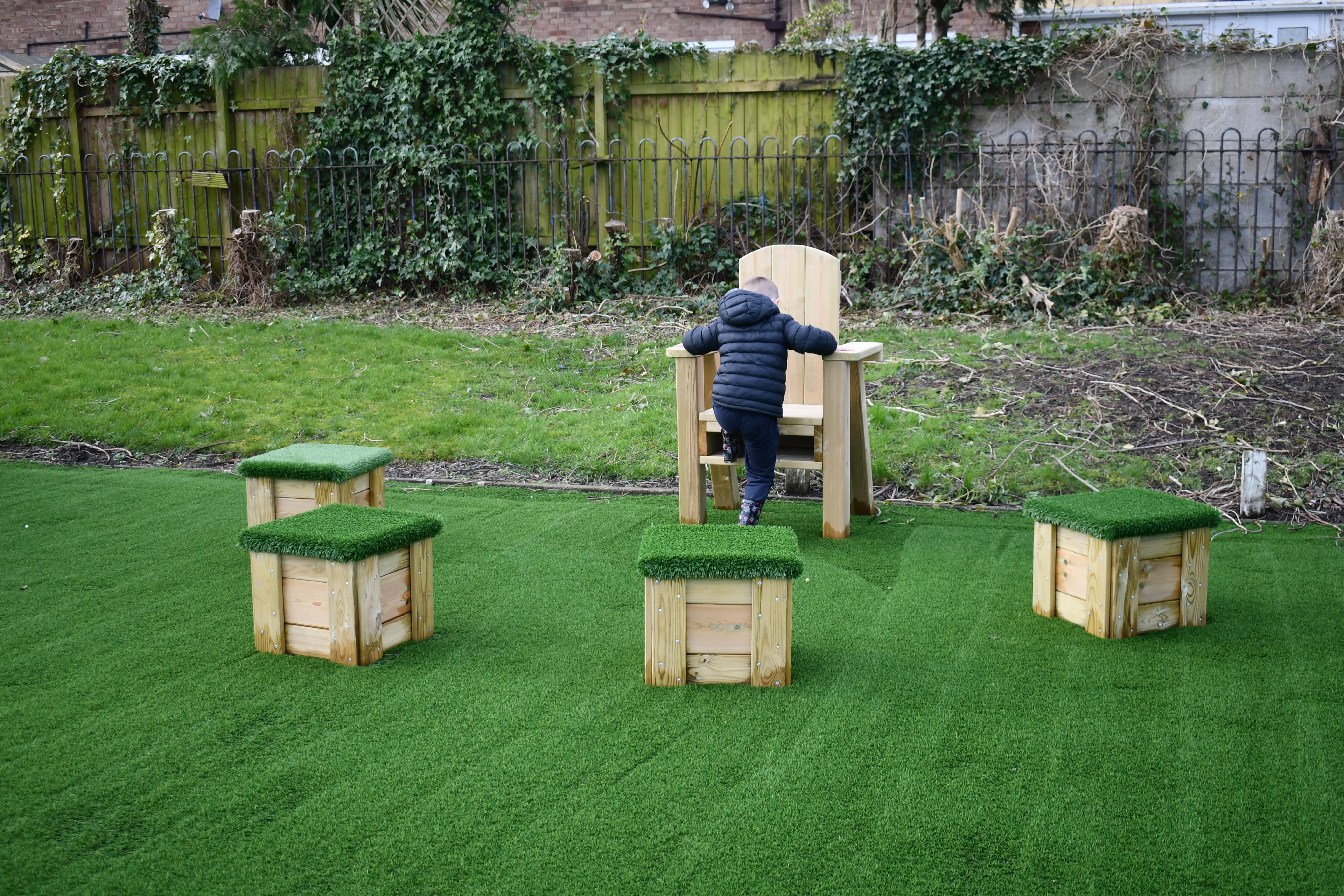 A child is climbing onto a big wooden chair, with 4 wooden blocks surrounding the chair. The wooden blocks are seats with artificial grass tops.