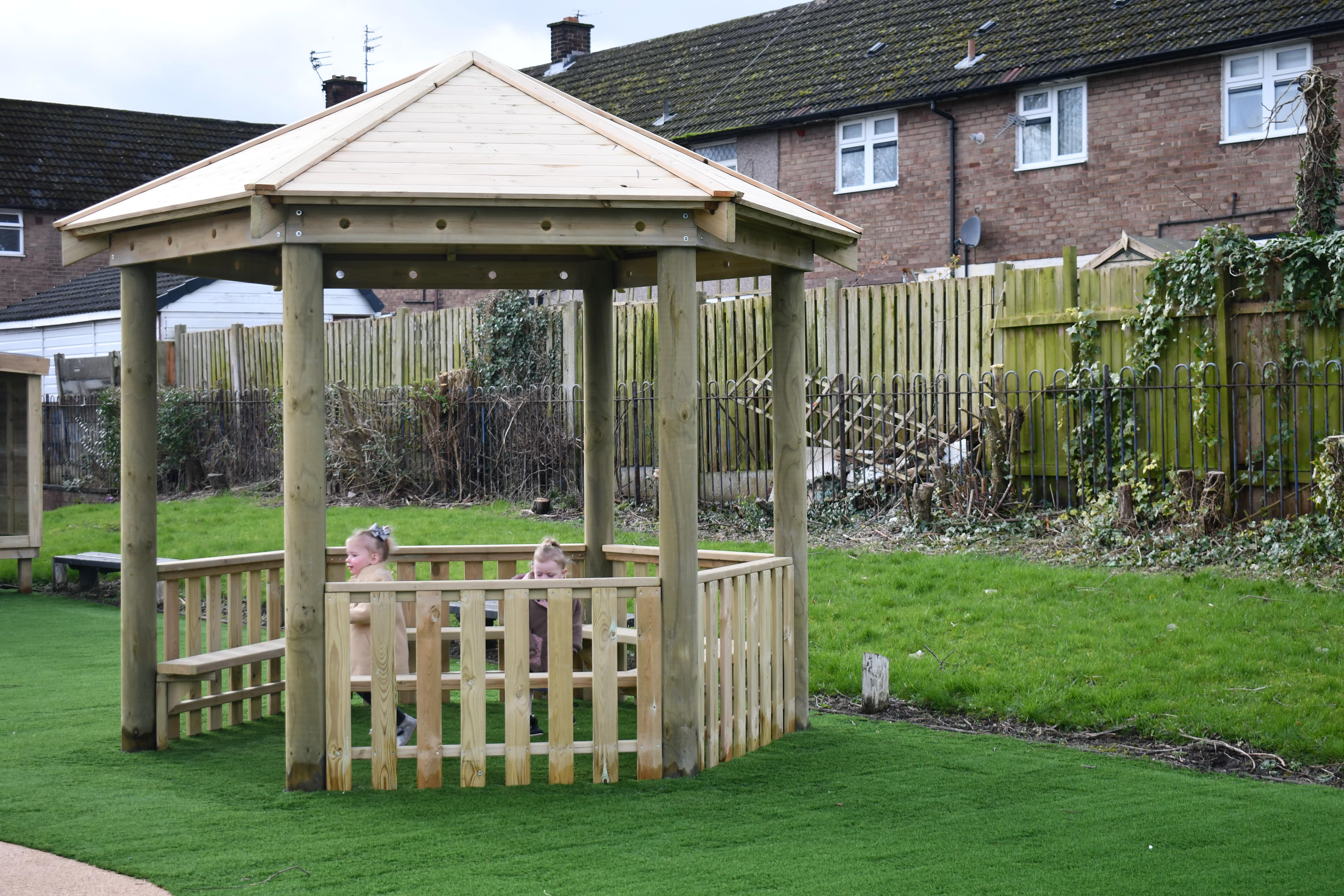 A wooden canopy has been installed on the artificial grass that has been placed on the play area. Two children are sat inside and are looking around.