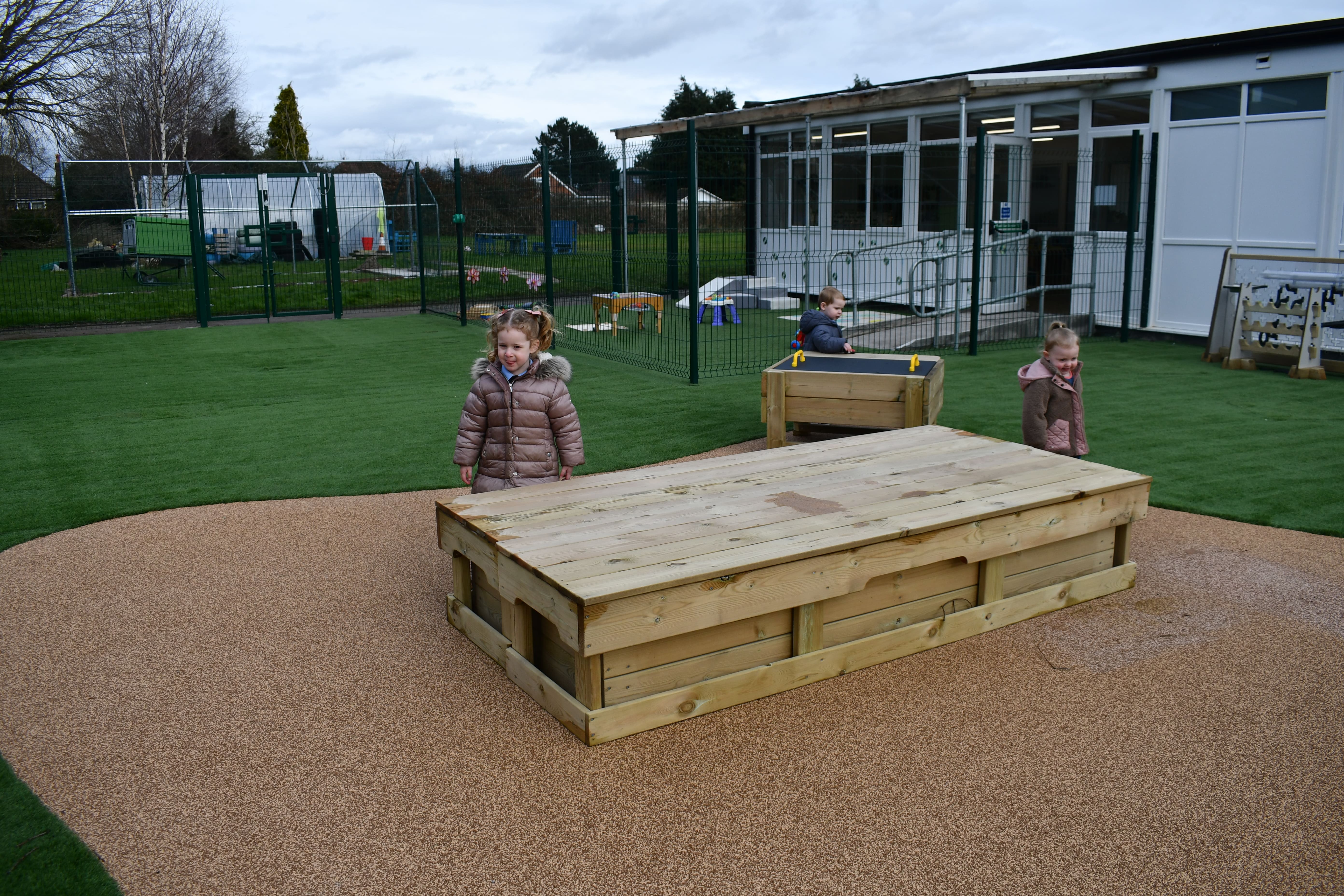 Two children are exploring the large sandbox and are walking around the structure. A child in the background can be seen engaging with another science station.