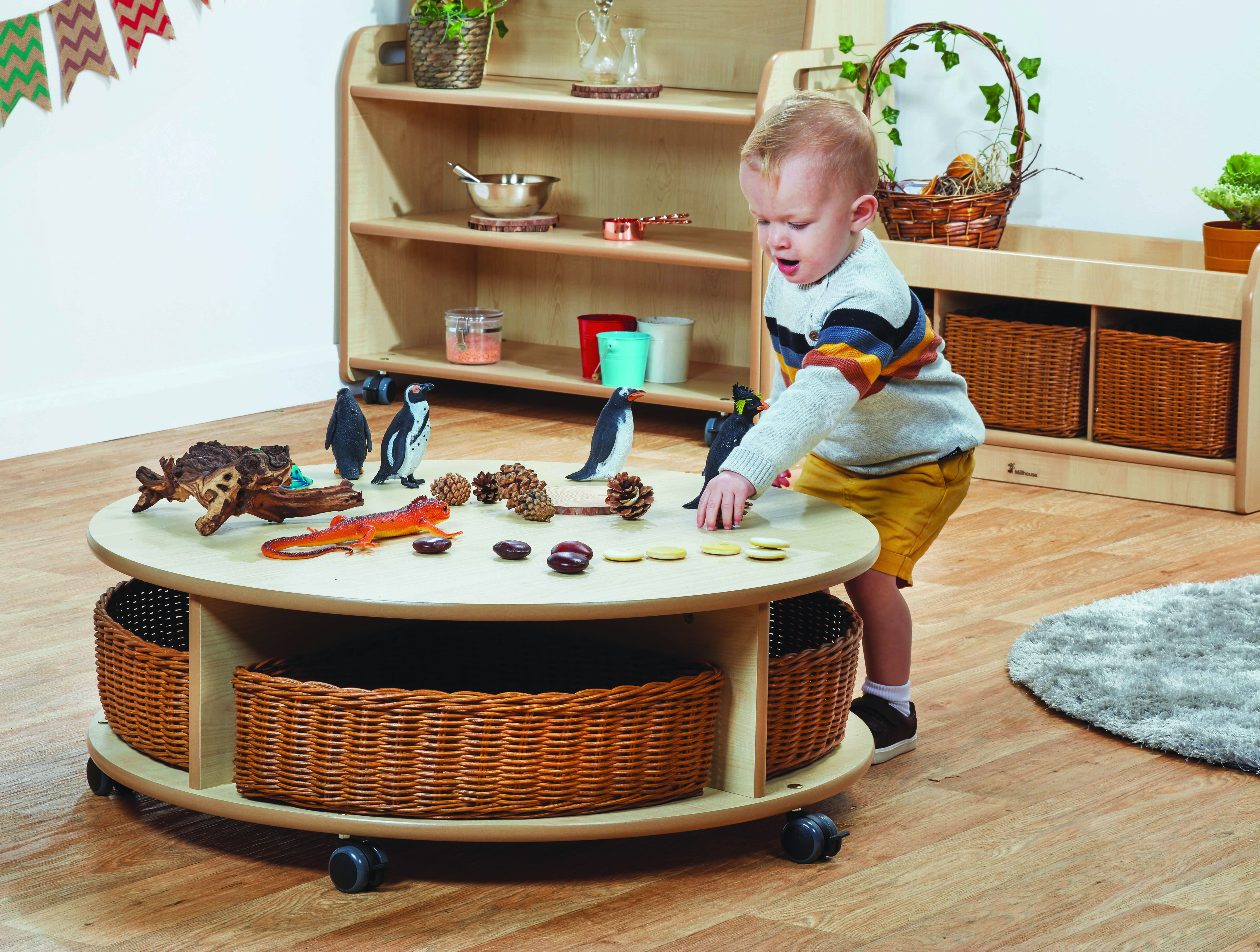 A boy is playing with toys on top of a small, circular storage unit. The storage unit has wicker baskets included and wheels can be seen at the bottom.