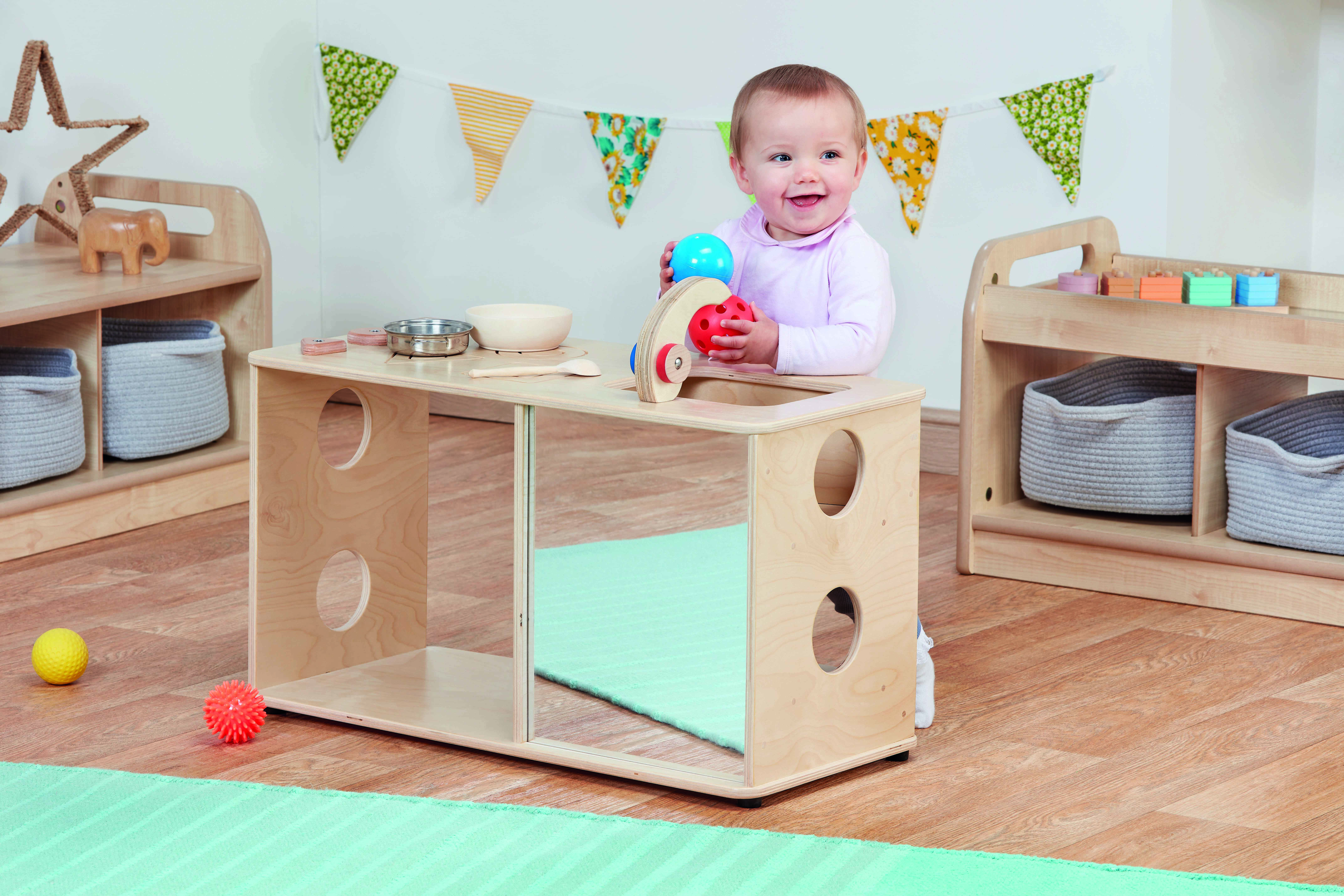 A toddler is playing with a simple role play kitchen set, and is holding the tap. The baby is looking off-camera and smiling.