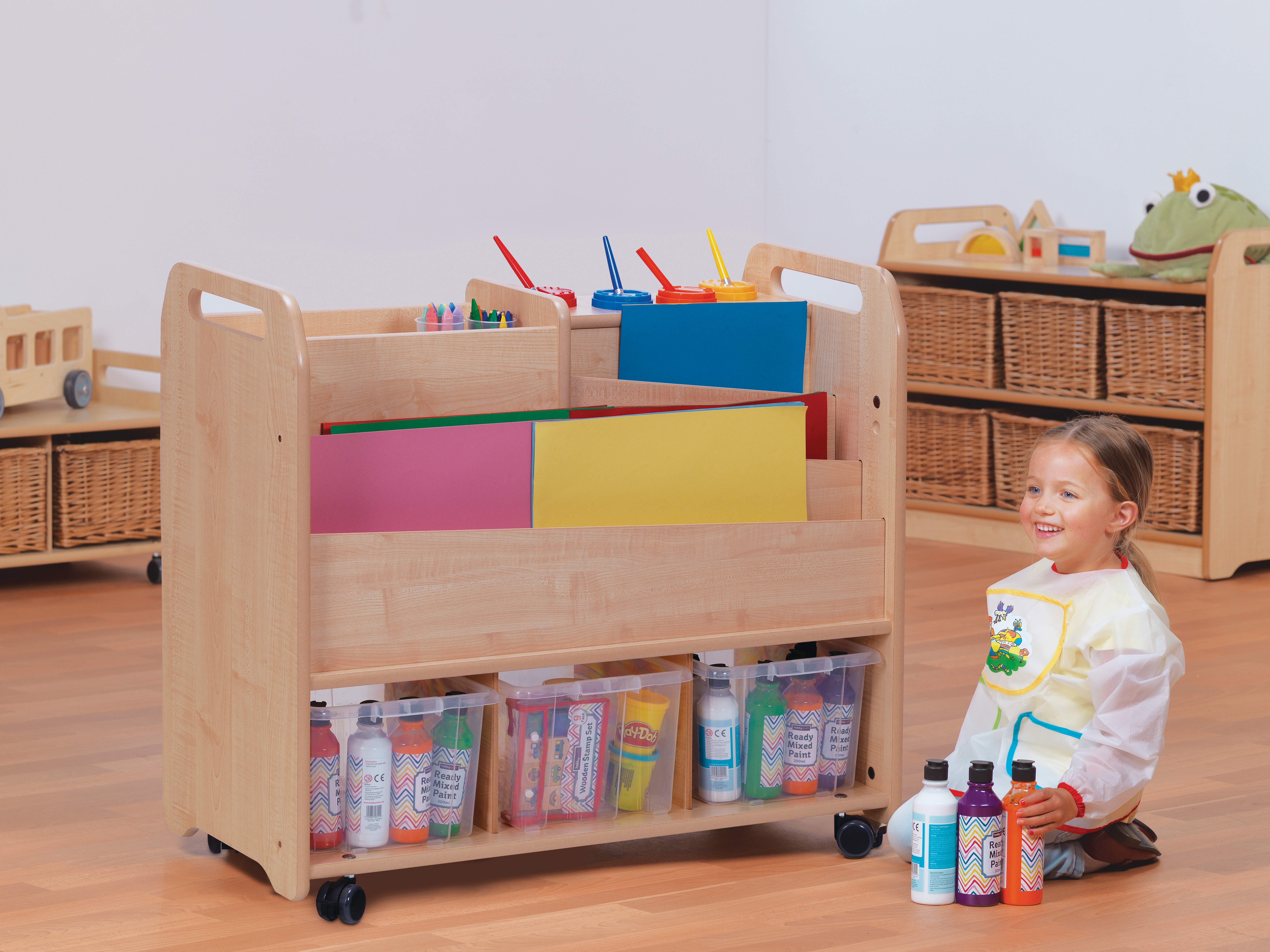 A little girl is sat on the floor beside an arts and crafts provision trolley. She has three bottles of paint placed next to her that are all different colours. She is smiling to someone off camera.