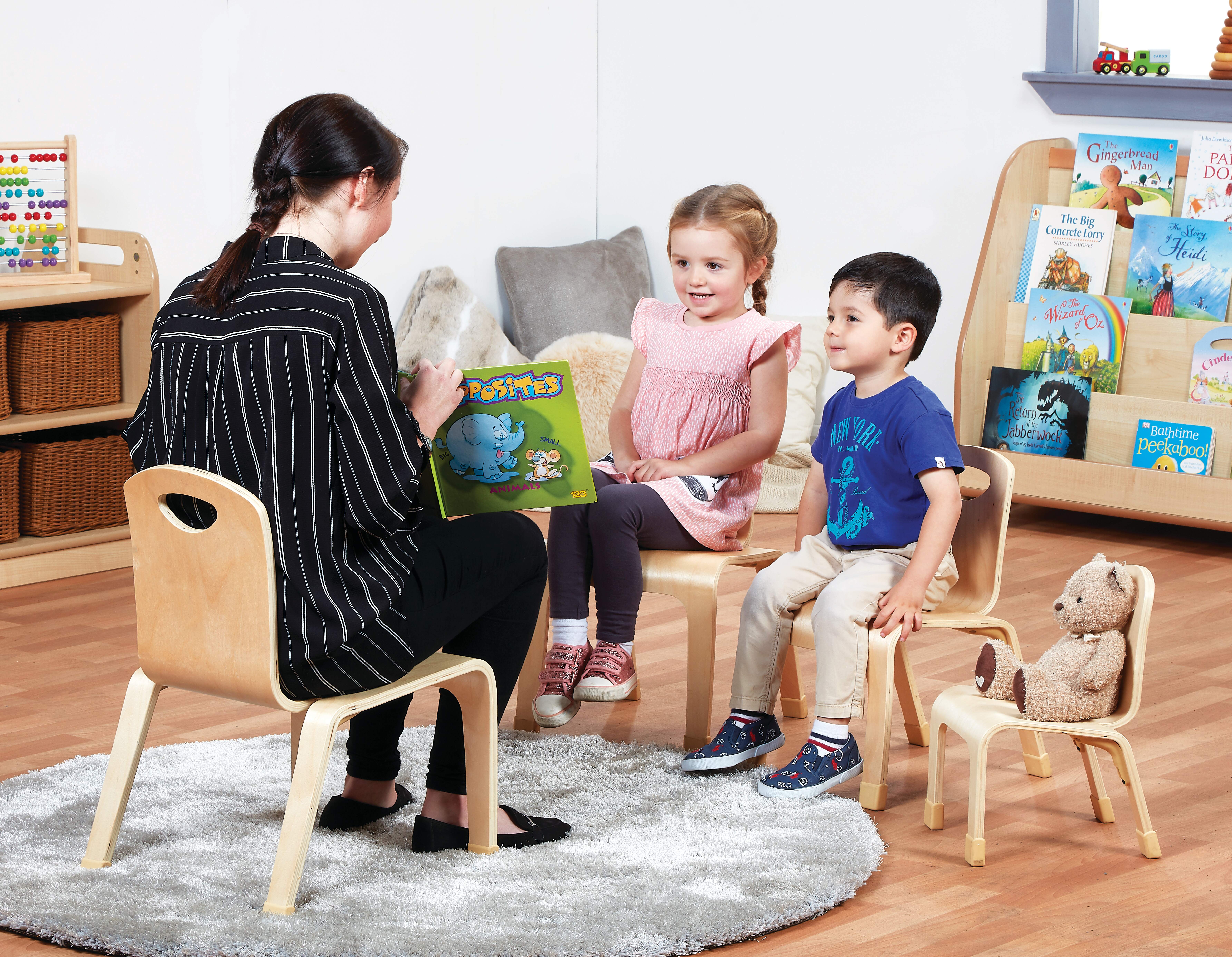 A teacher is reading a book to her kids. The teacher is sat on a big wooden chair as two children sit opposite on wooden chairs. A teddy is placed beside the second child on a wooden chair.