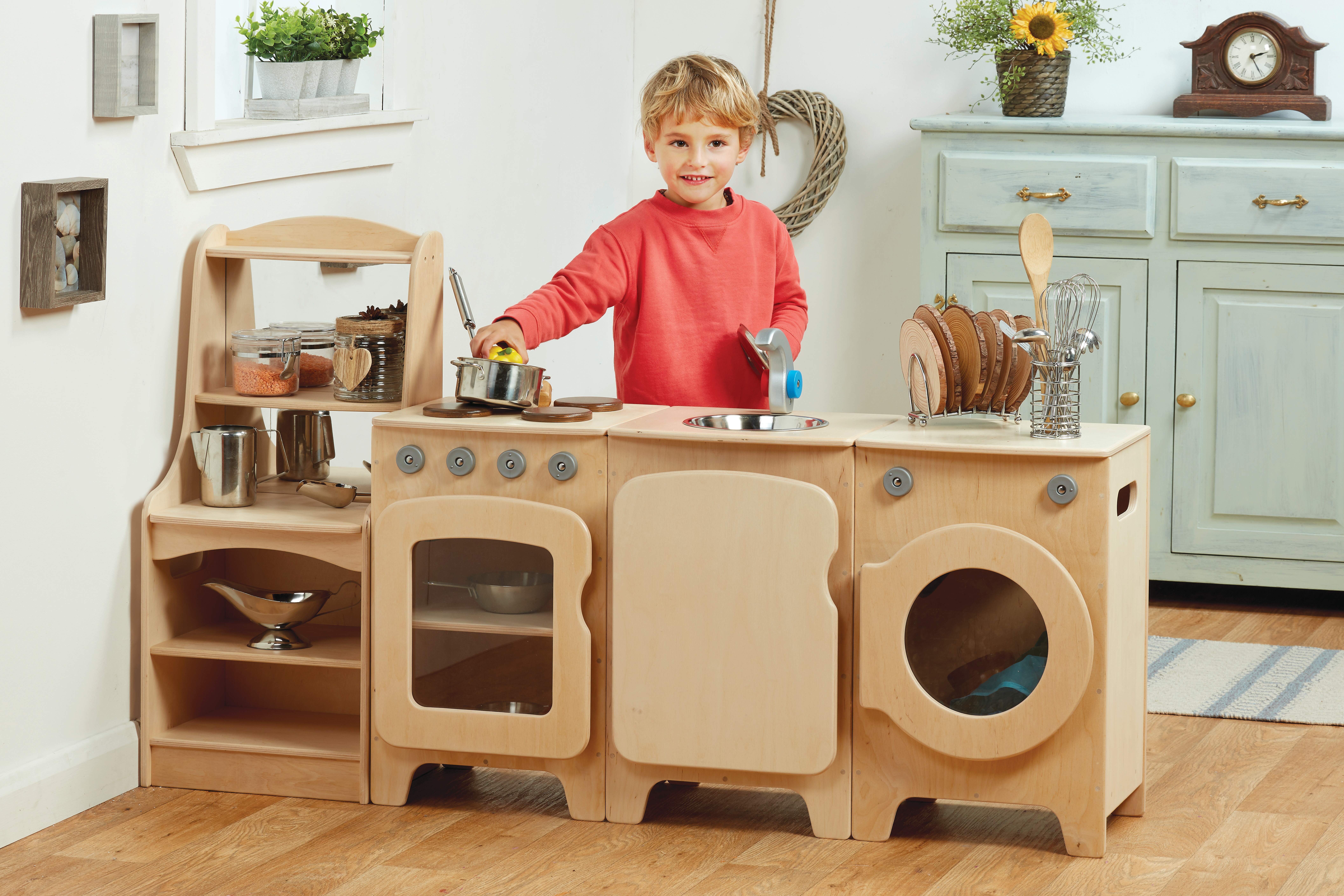 A boy is playing with a big role play kitchen and is holding a pan over the stove. The kitchen has a storage unit, an oven, a dishwasher and a washing machine, all attached to each other.