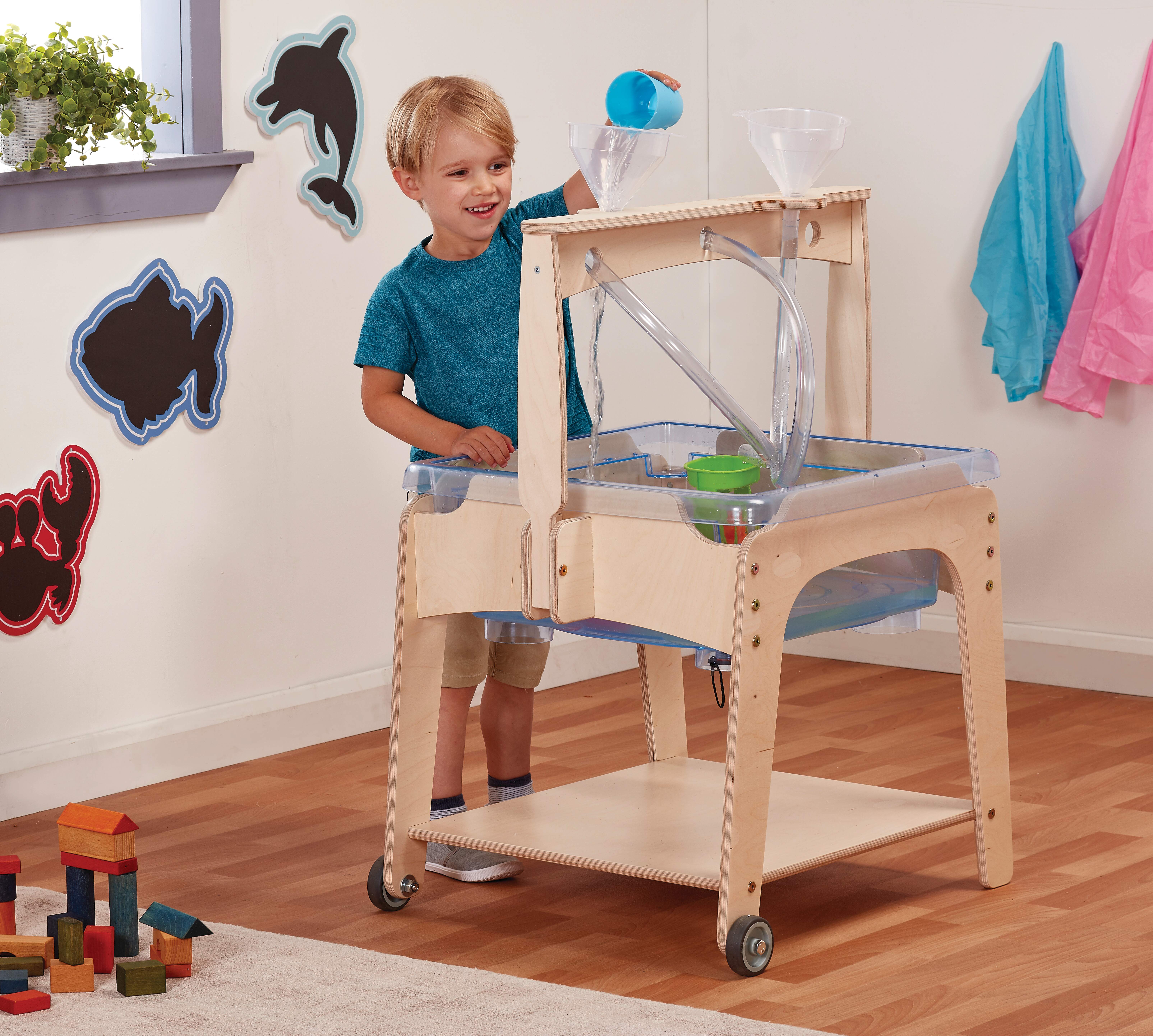 A little boy is pouring water down a funnel, which connects to a clear pipe that leads to a plastic tub. The tub is supported by a wooden structure.