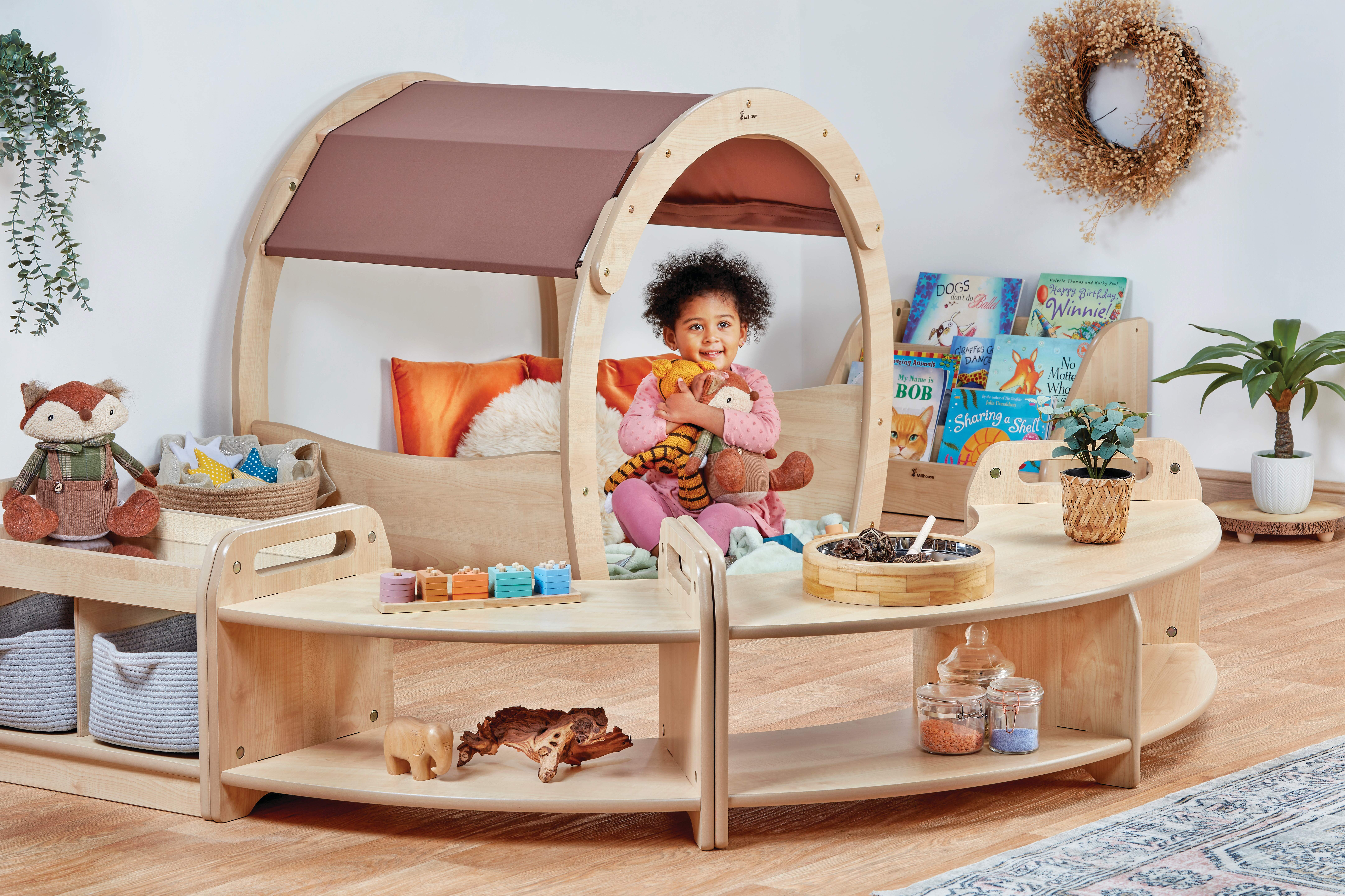 A little girl is sat in a wooden den, with pillows and teddies placed inside. A set of low angled shelves are placed around the den, creating a zoned off area.