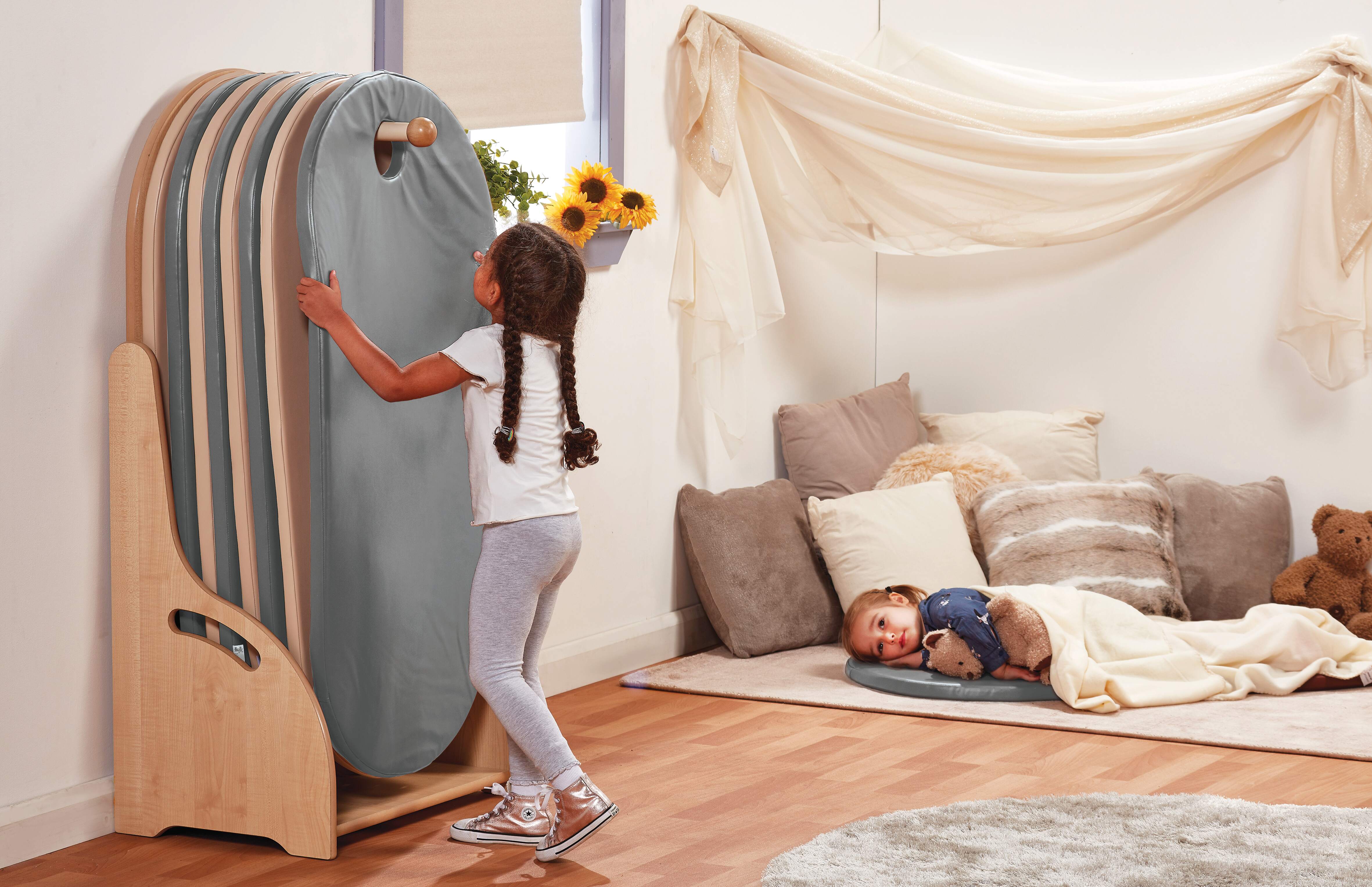 A girl is collecting a sleeping mat from a mat storage unit. The mat is grey and beige. Another girl can be seen falling asleep on a mat that is next to the unit, surrounded by pillows.