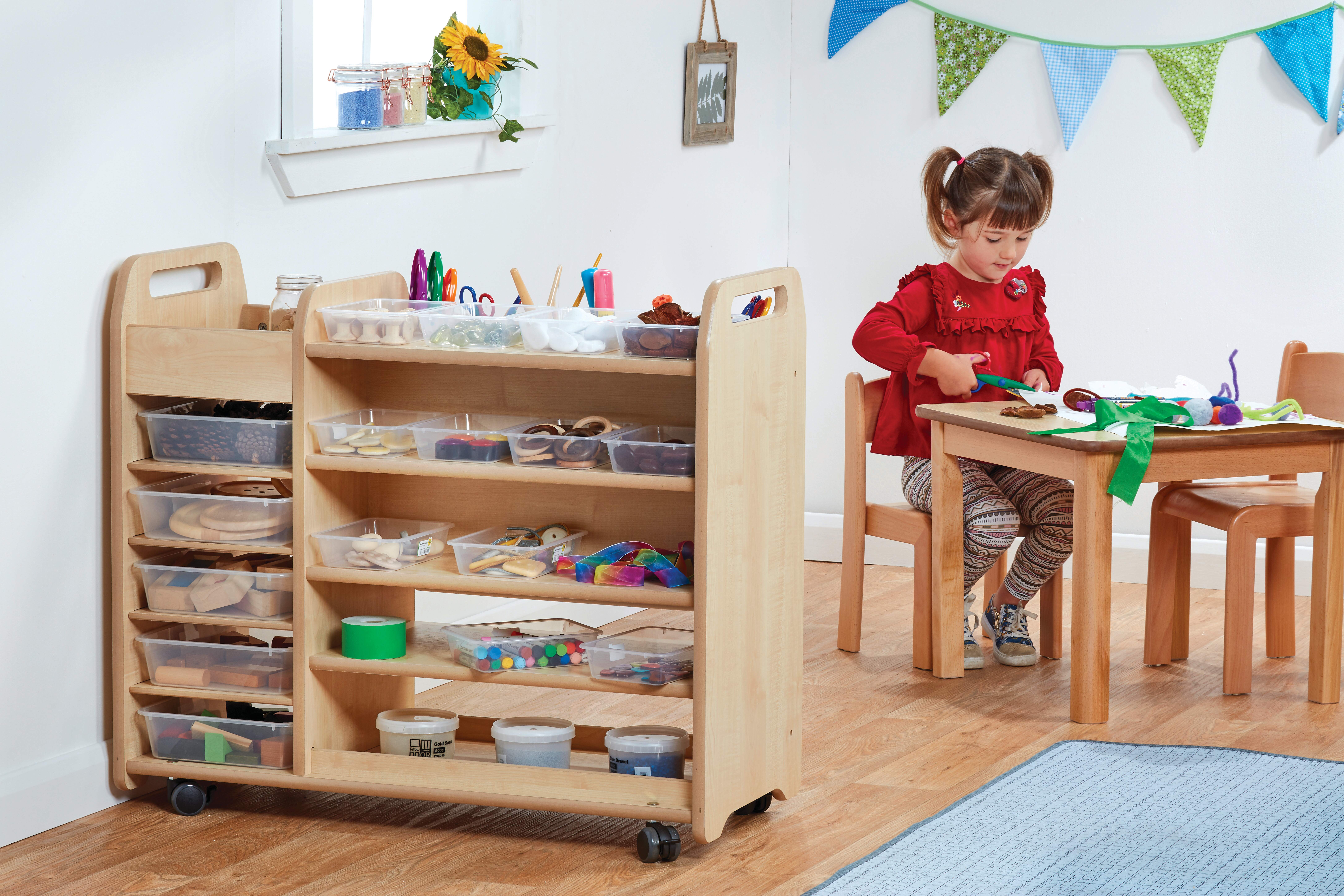 A little girl is sat at a table as she cuts up some paper. Next to her is a wooden trolley with wheels which is holding a wide variety of different arts and crafts pieces.