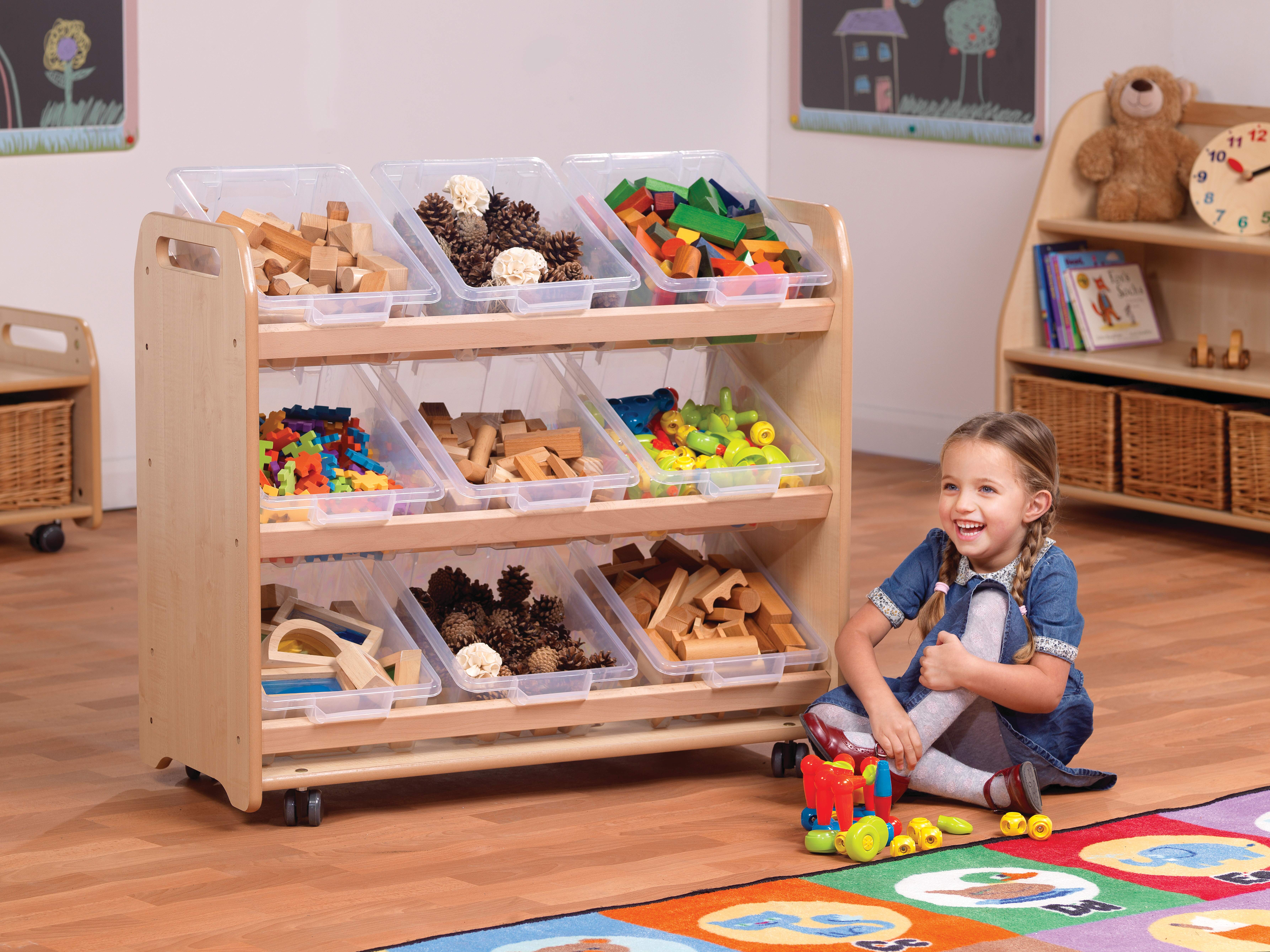 A little girl is sat on the floor next to an inclined storage unit on wheels. Each of the 9 boxes on the storage unit hold different toys in them.