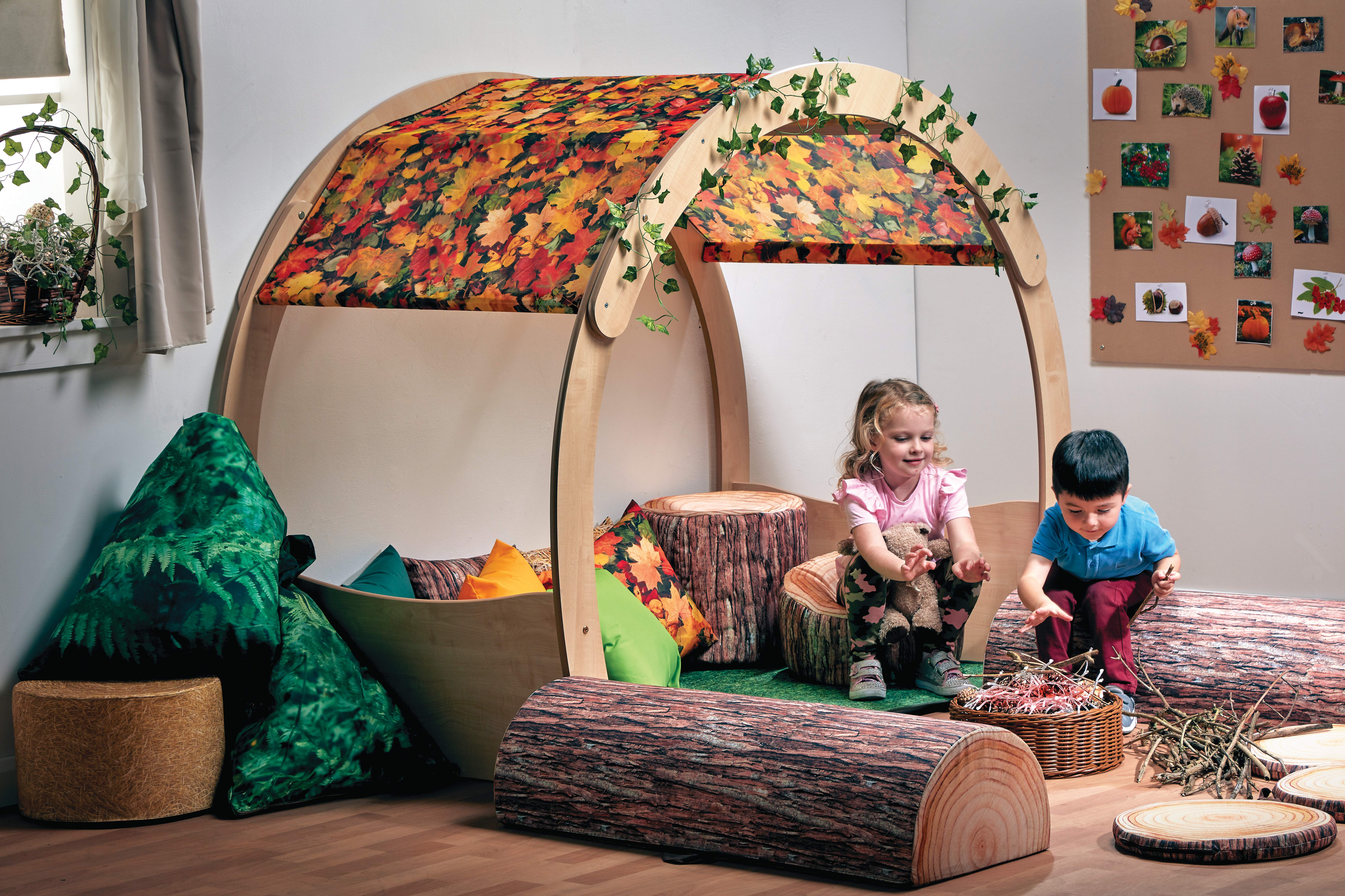 Two children are sat on logs as a wooden indoor den covers them. It has an autumn print roof and has a variety of autumn coloured pillows and toys inside of the den