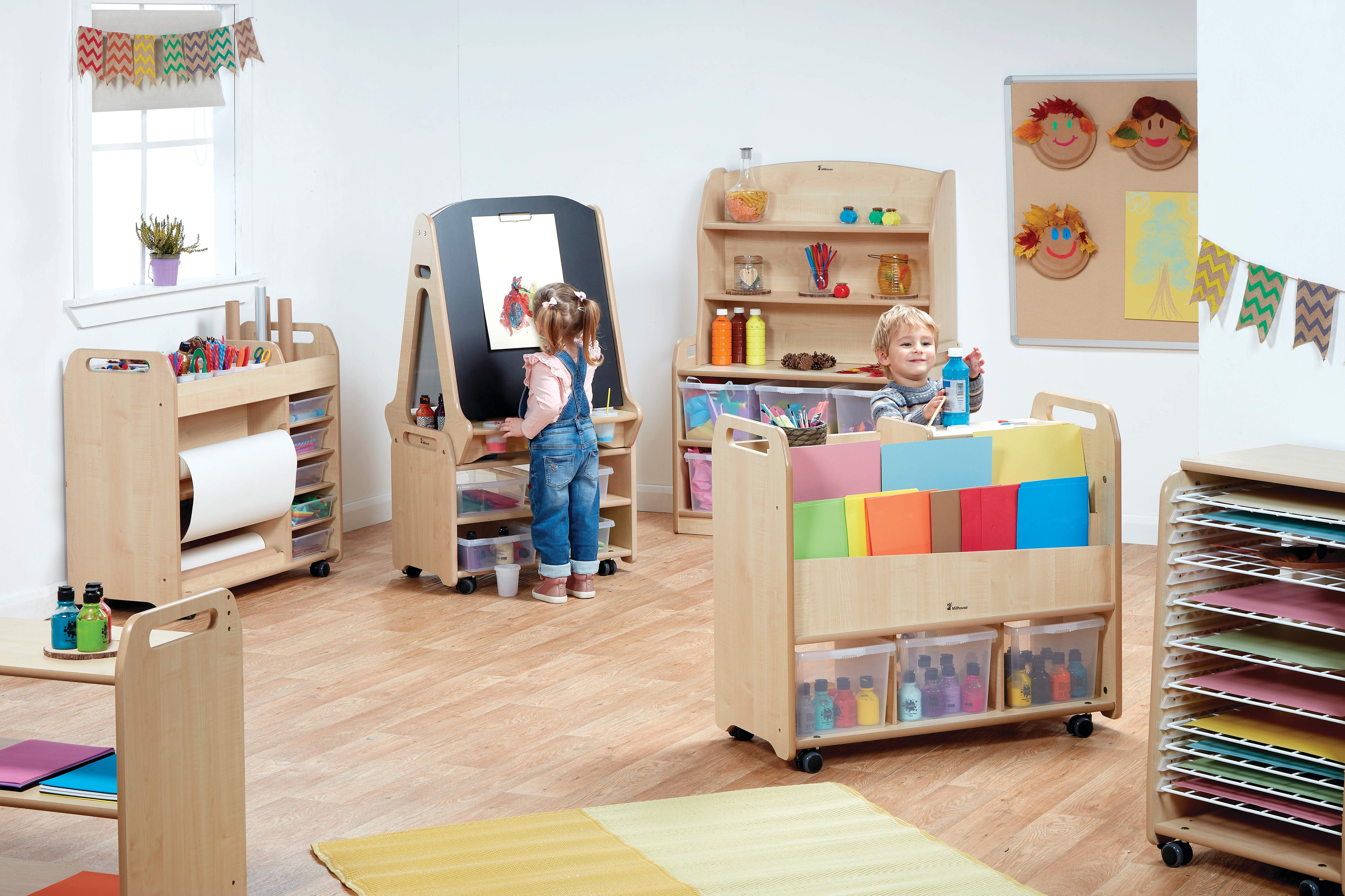 Two children are in a room that is full of different arts and crafts furniture. A girl is drawing on a chalkboard on wheels whilst a boy is grabbing paint for an Arts and Crafts provision trolley