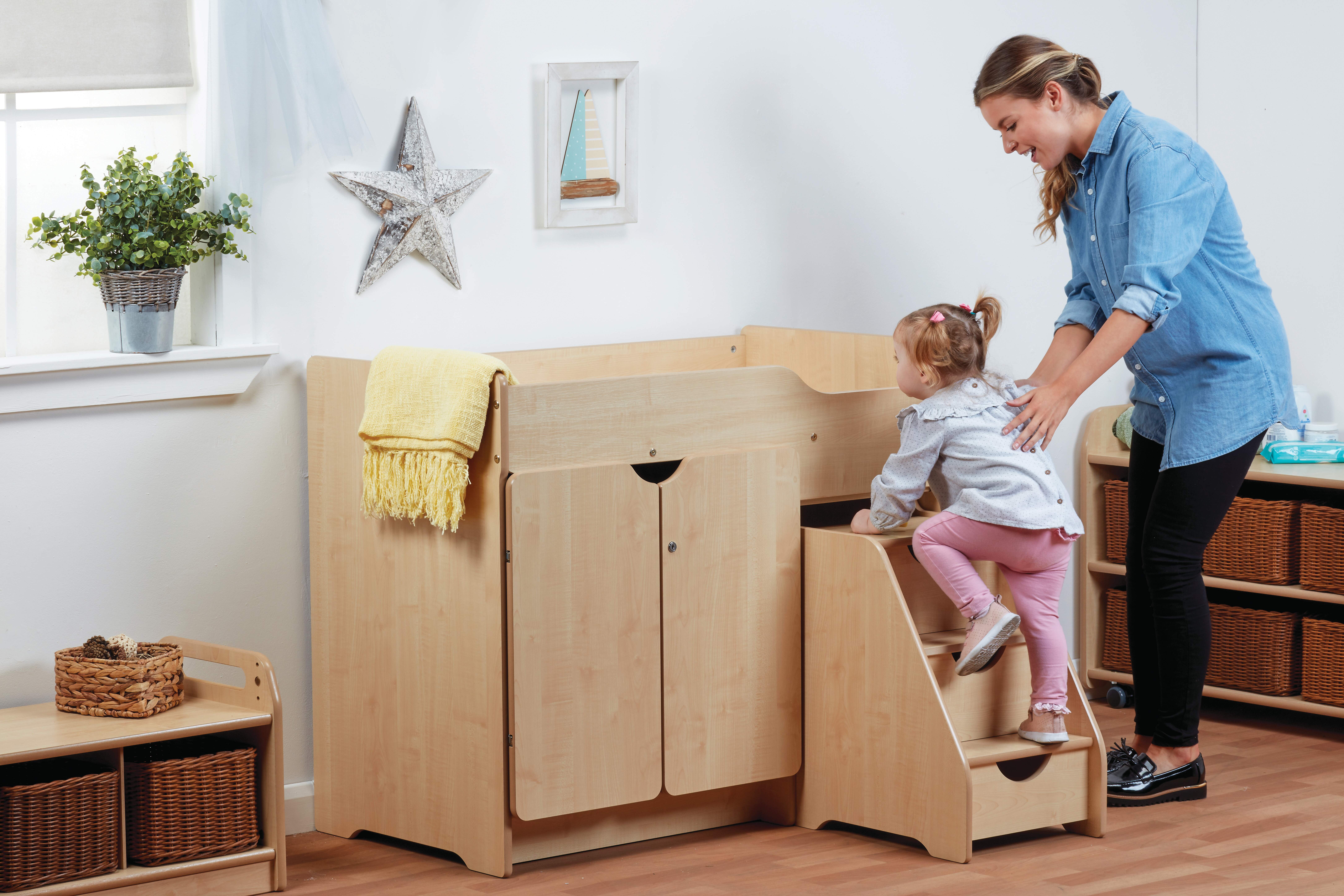 A toddler is climbing up some steps with the assistance of an adult. Above the steps is a changing unit, with a yellow blanket hanging over the side.