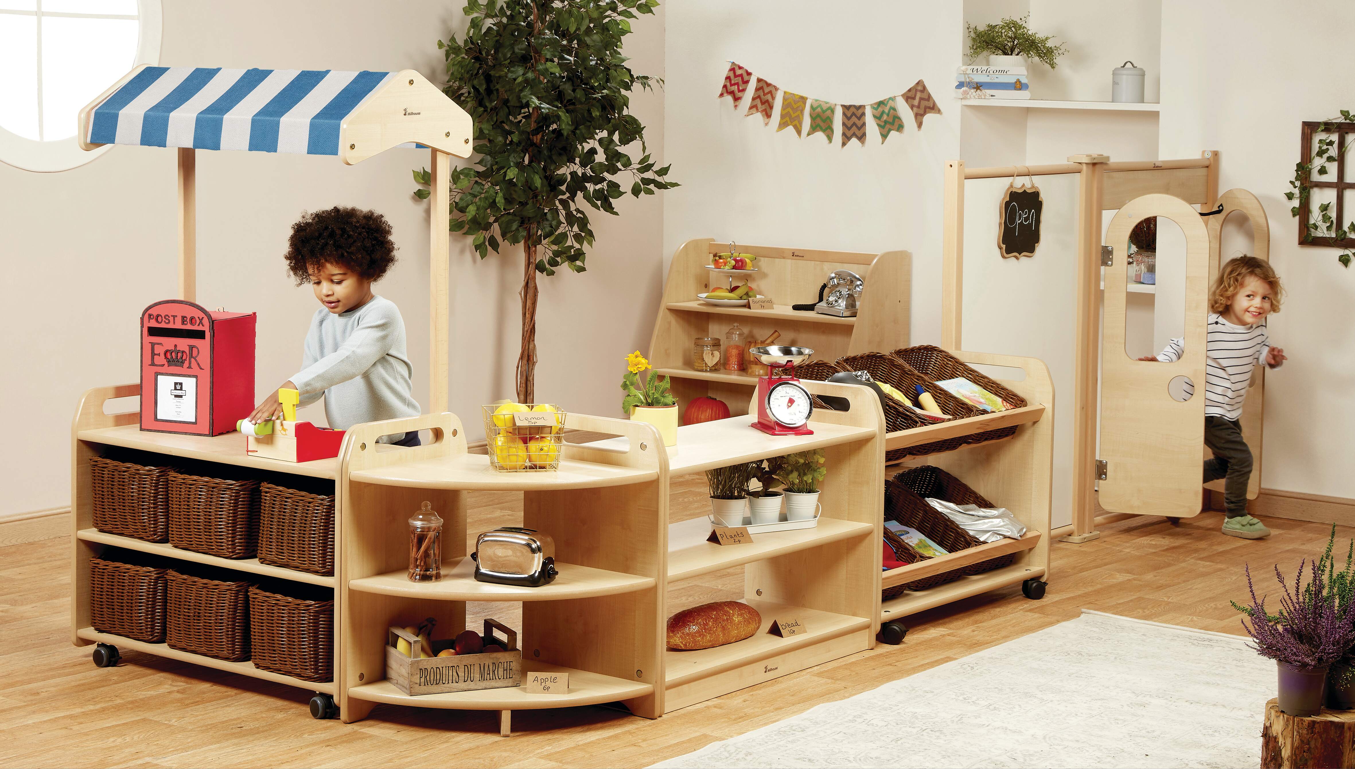 A little boy is stood behind a role play market stall as he operates a toy cash register. Connected to the stall are some shelves showcasing different toy props. Another little boy is entering the room through a little wooden door.