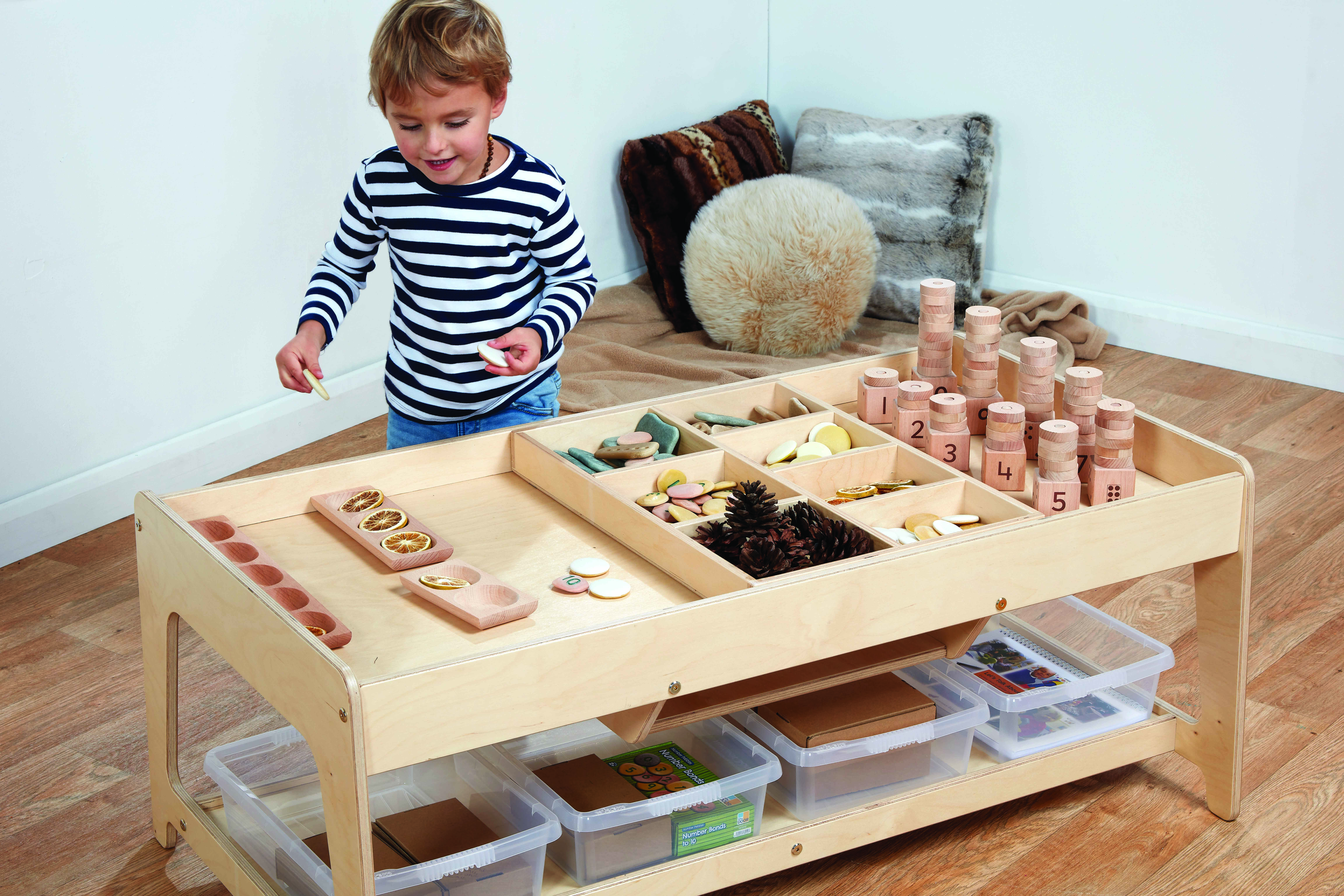 A boy is playing with a variety of props on a wooden table. The table has two levels. The first level contains clear tubs to store objects in and the second level is split into subsections, promoting investigative play.