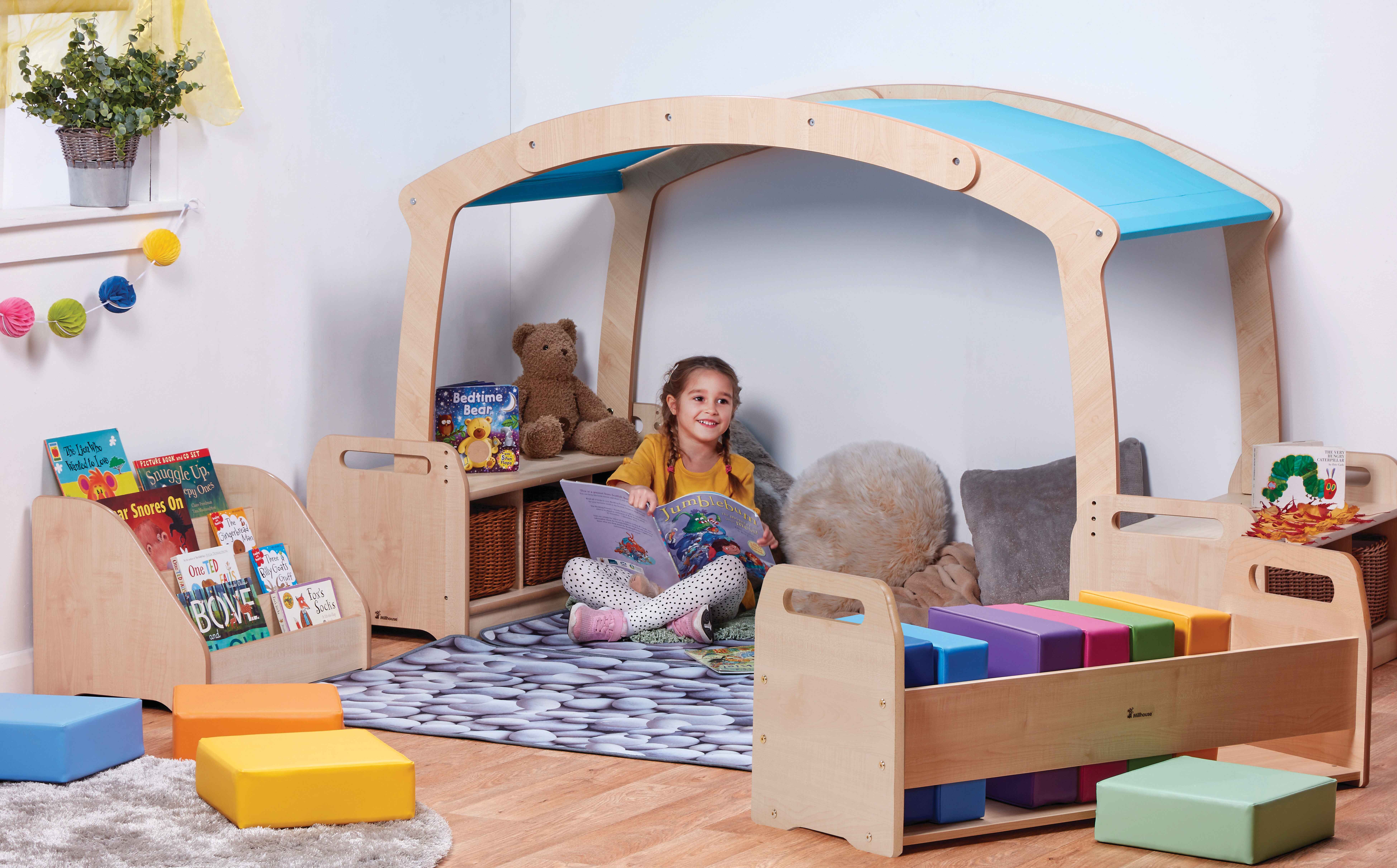 A child is sat underneath a small, indoor wooden canopy that has a blue roof. The girl is reading a book as other pieces of furniture surround her.