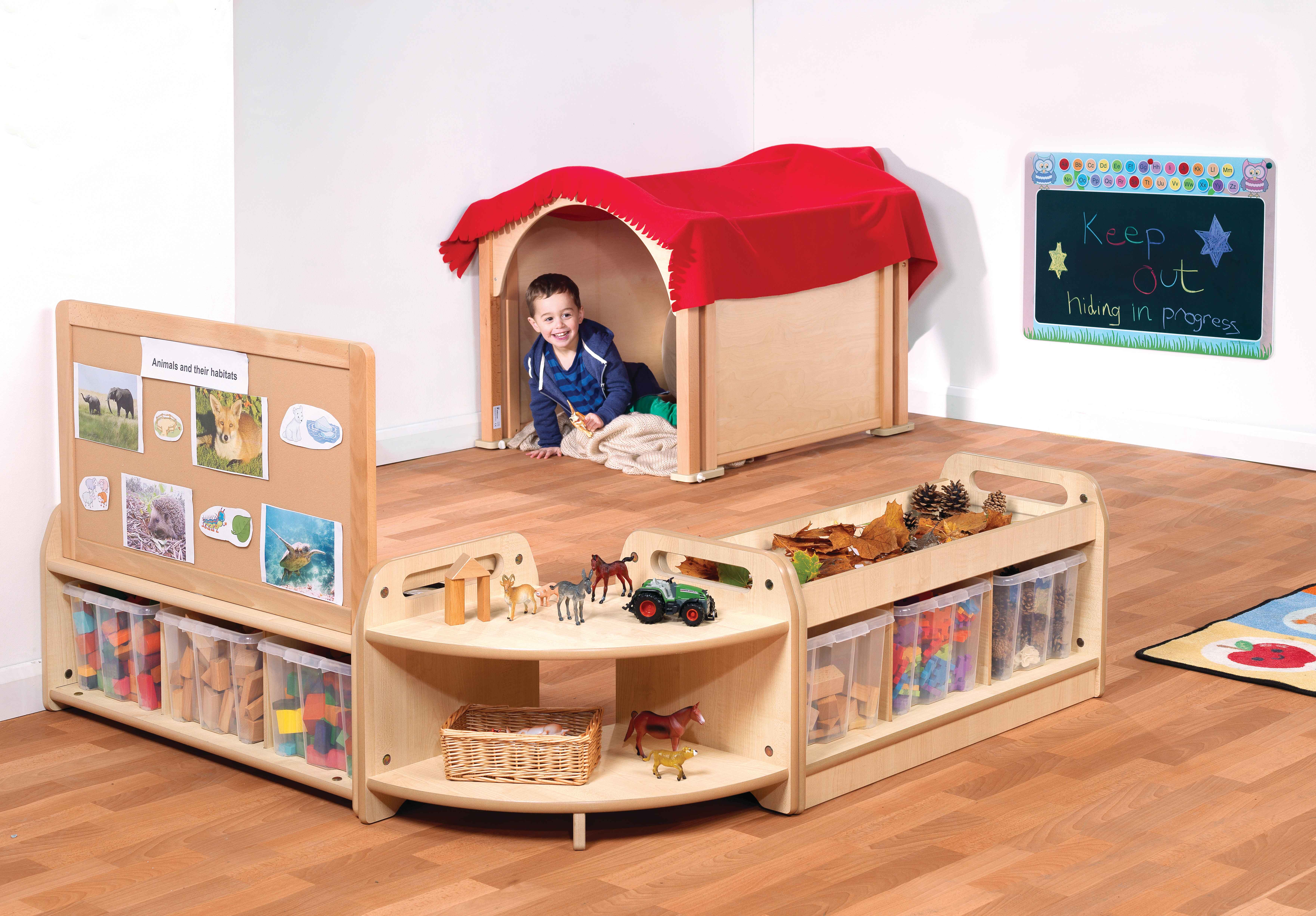 A child is in a wooden den, with a red blanket covering the roof. A set of shelves and a mobile storage unit has been placed nearby, creating a zone