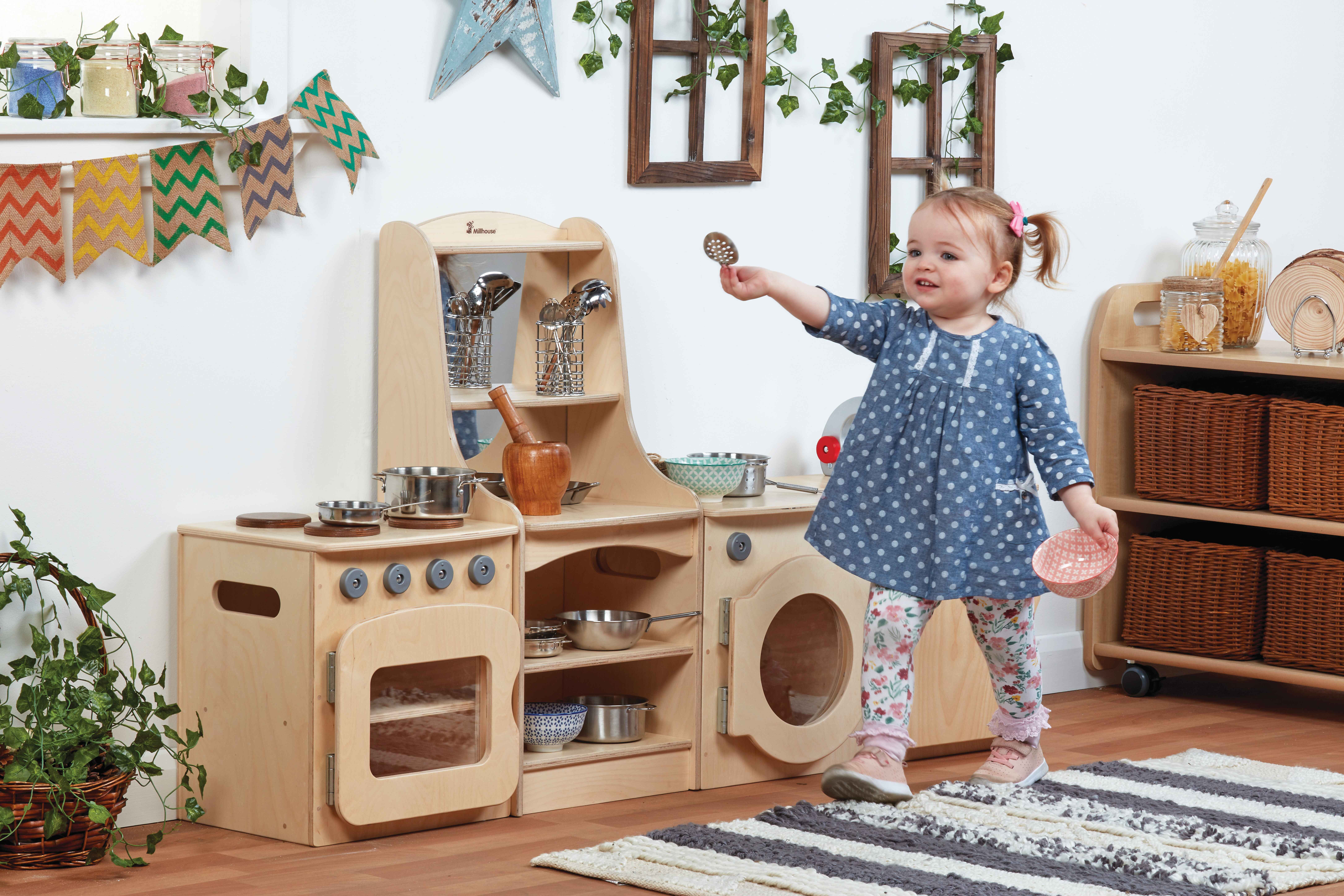 A little girl is stood next to a play kitchen set and is holding a metallic spoon in the air