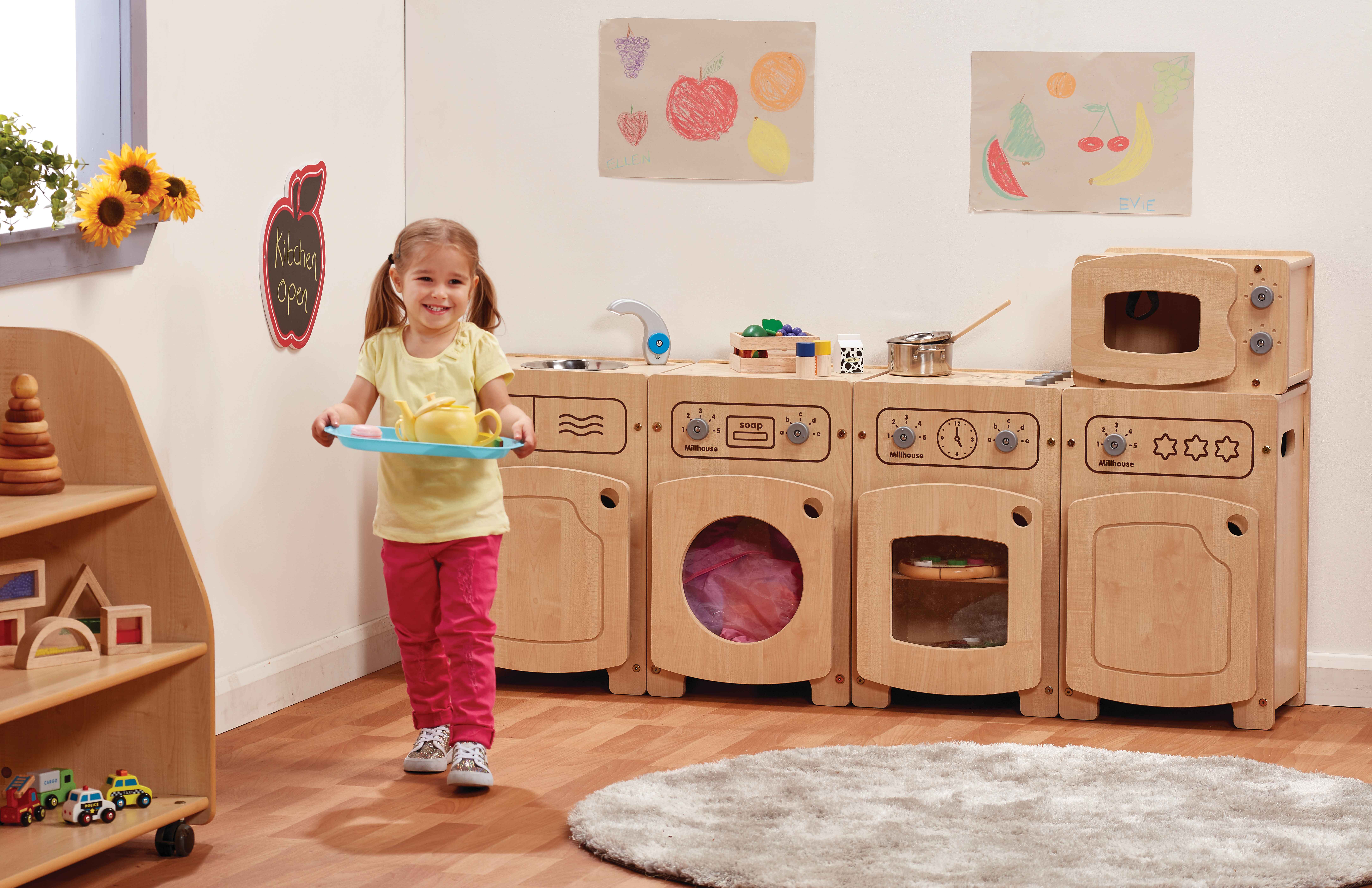 A little girl is holding a tray that has a toy kettle placed on it. She is walking towards the camera and away from a kitchen role play set.