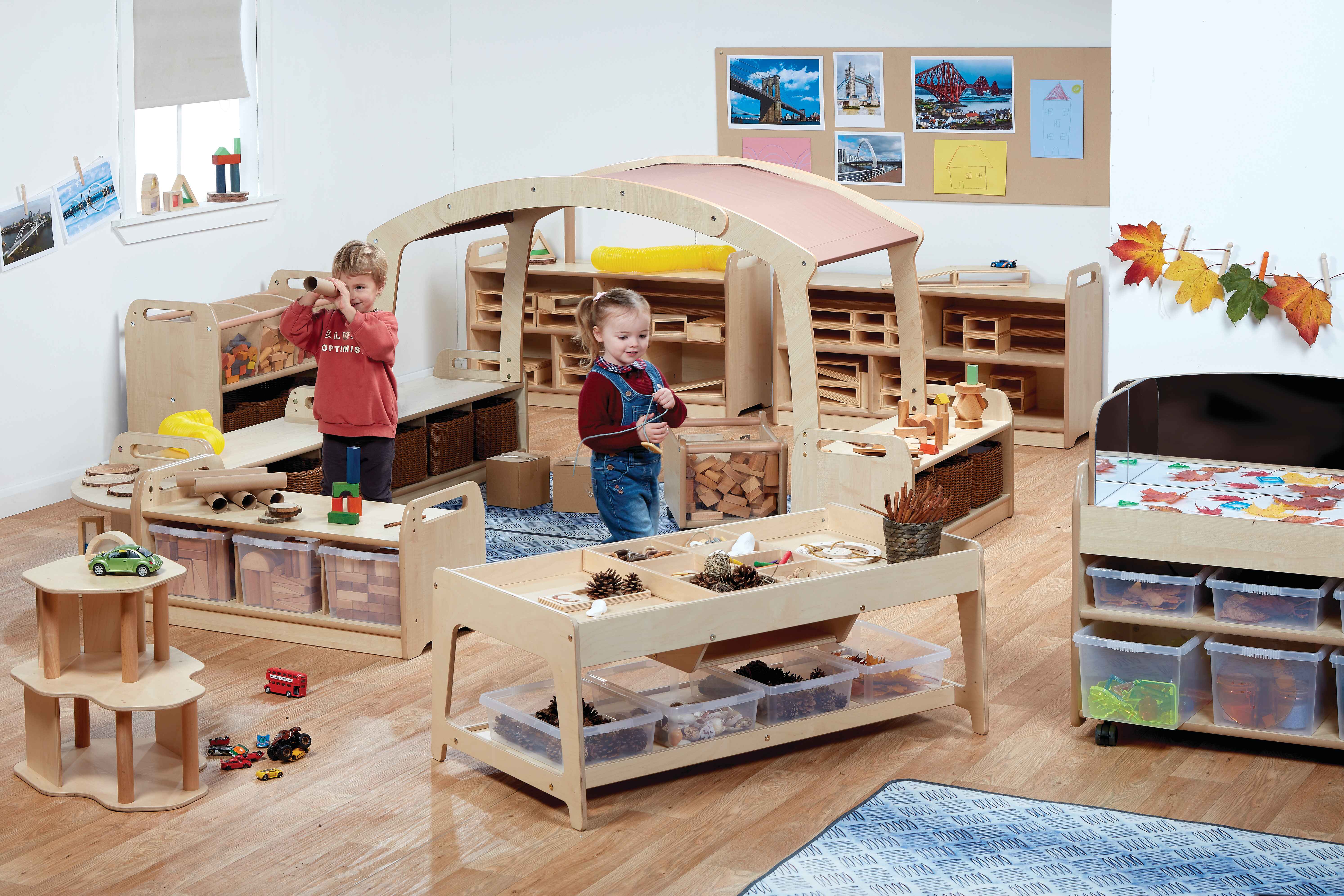 Two children are exploring a construction play area. A variety of construction play equipment has been placed around the room, from storage unit containing STEM blocks to construction tables. One child is holding up a cardboard tube and is using it as a telescope whilst the other child is approaching a construction table.