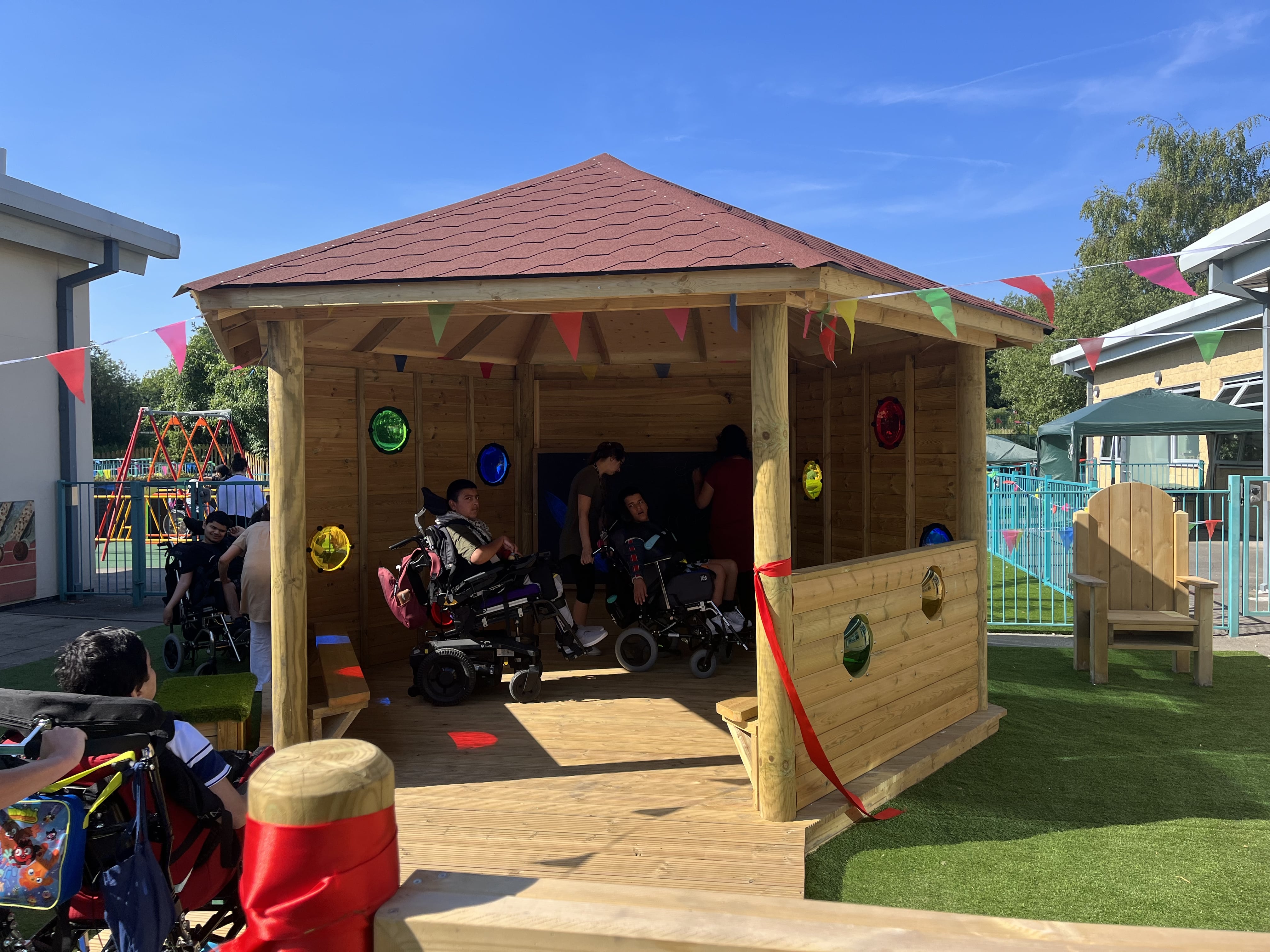 A wooden gazebo that has had sensory features built into the main structure. Children in wheelchairs are inside and exploring the new environment.