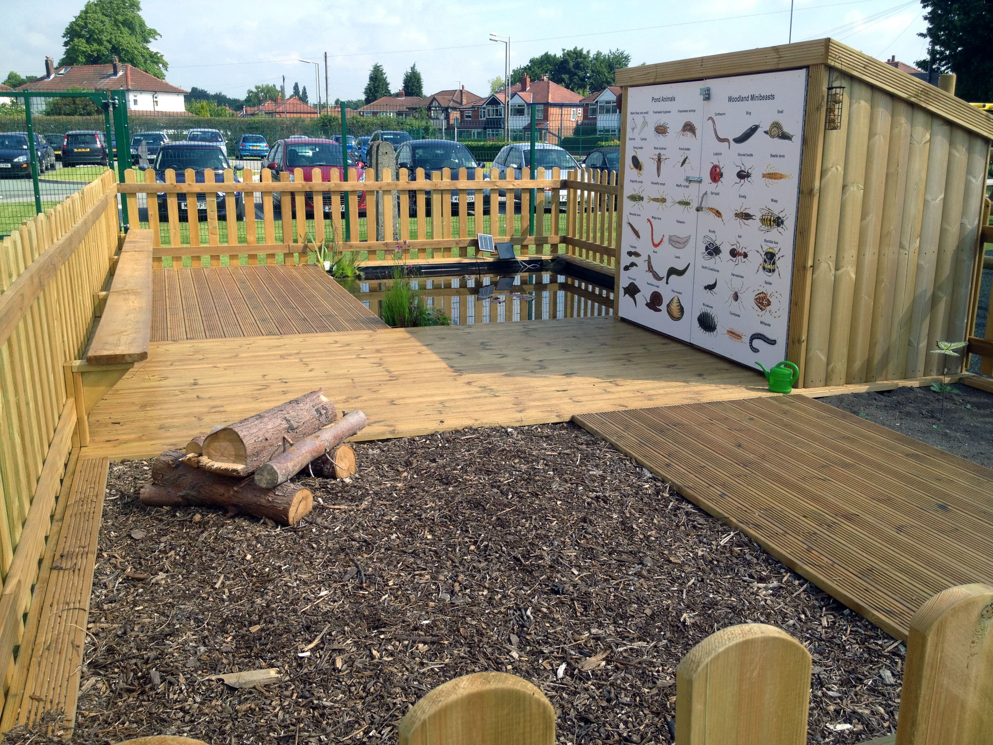 A wildlife area has been created with wooden pathways with a pond and a soil area present. A big structure can be seen with a board listing a variety of insects and amphibians. A wooden fence secludes the area.