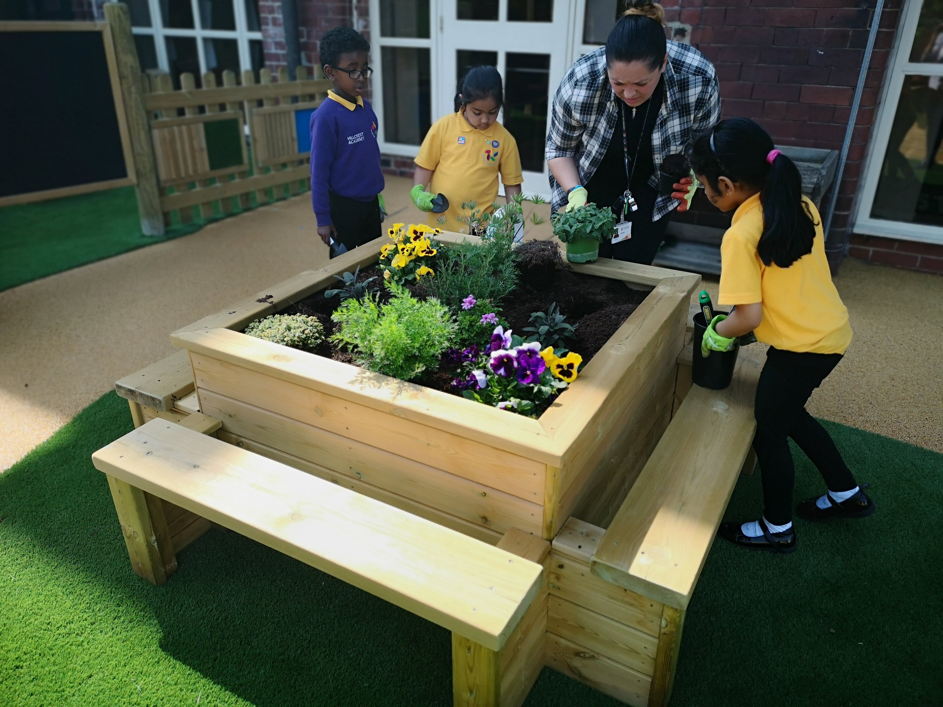 Three children and a teacher are all tending to a seated planter, which has some plants growing inside it. The children are holding different planting equipment as they tend to the plants.