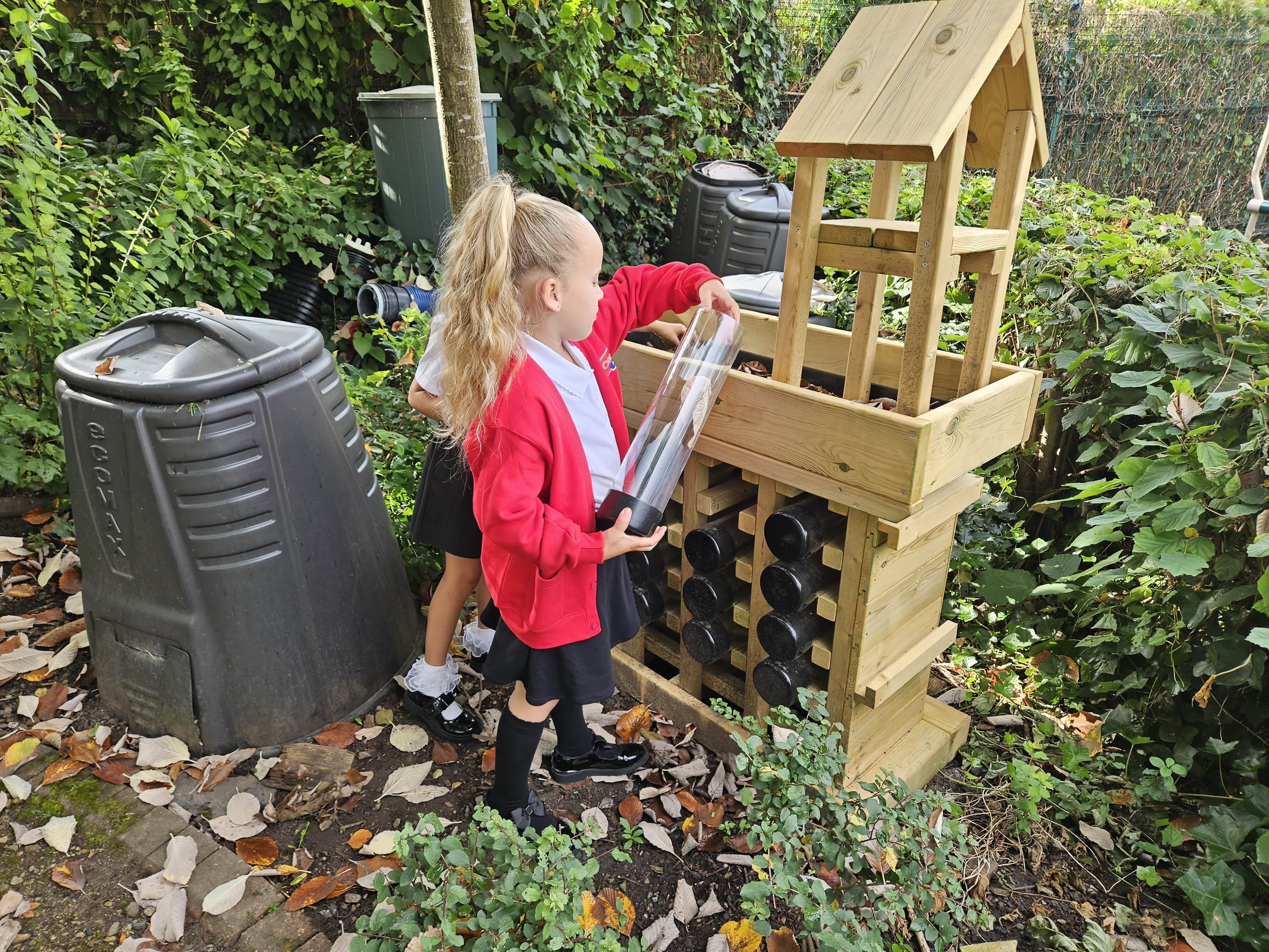 A child is holding a clear tube from a bug hotel wooden structure. The child is placing something inside the tube as another child is reaching for another tube from the wooden structure.