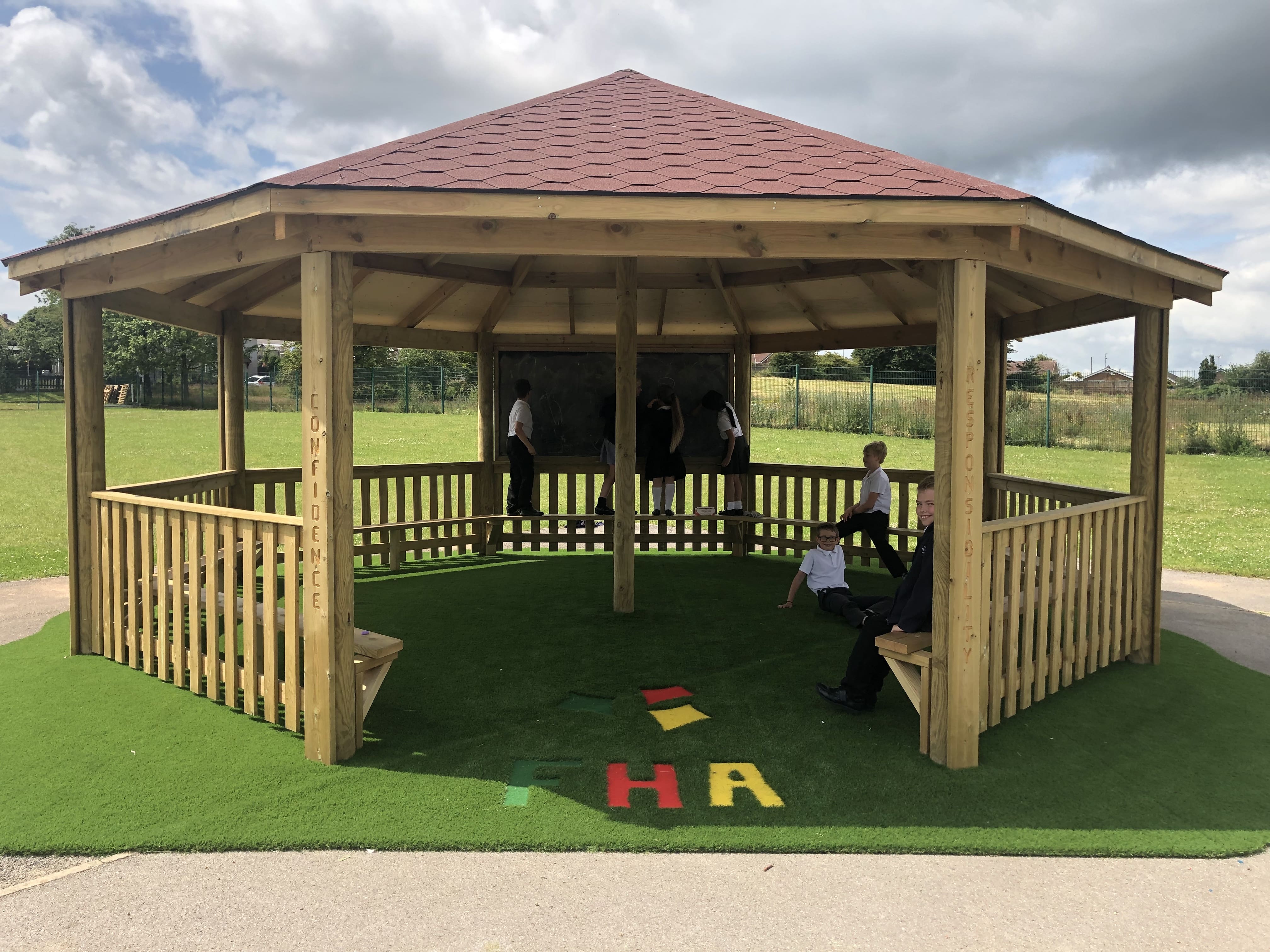 A big outdoor wooden gazebo that has a group of children underneath it, playing and sitting around. The flooring is artificial grass.