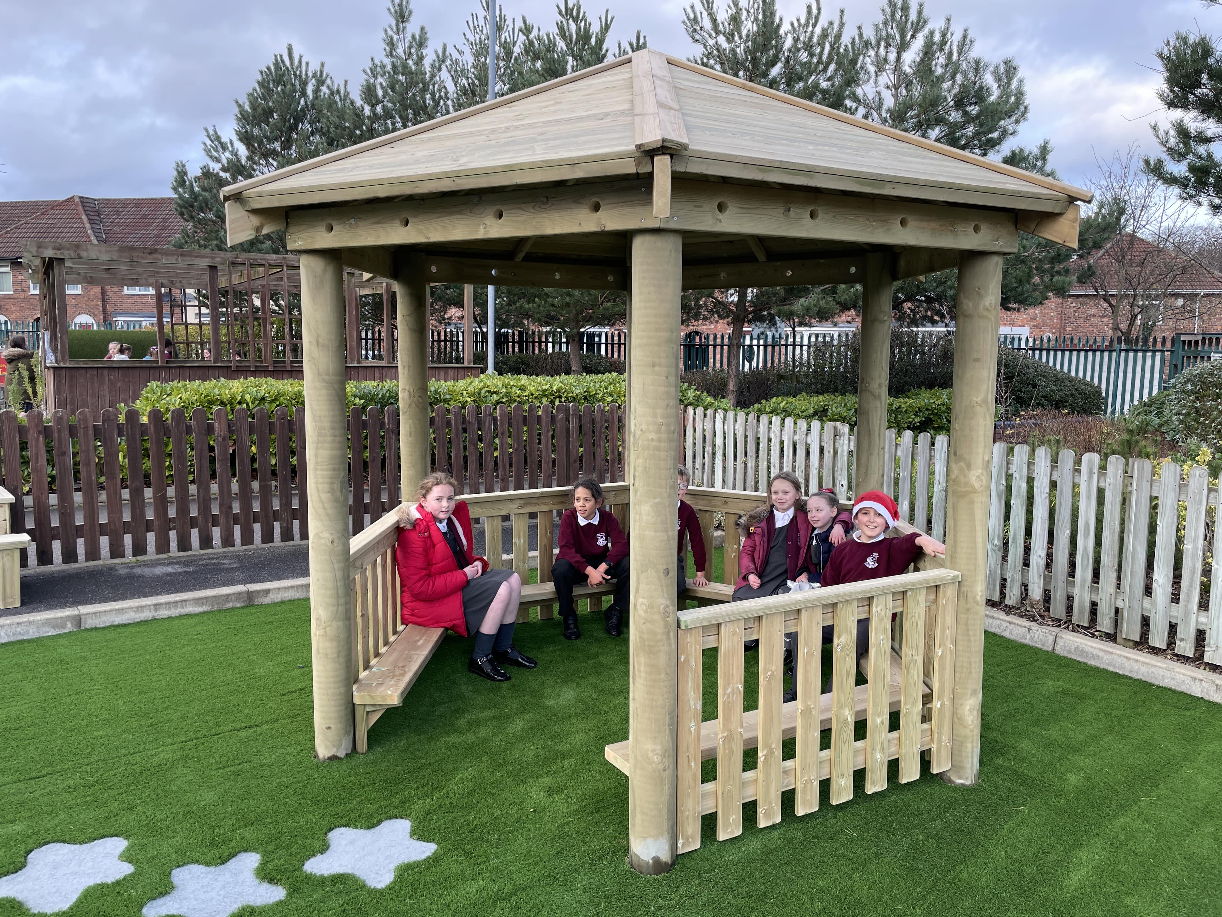 A 3.5m wooden gazebo with a group of children sat on benches that are built into the structure. The gazebo has been built on top of an artificial grass surface.