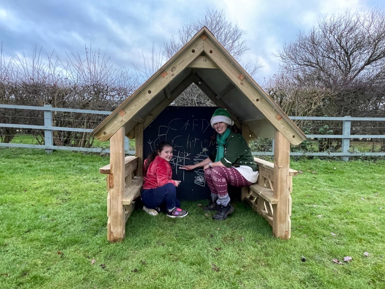 A member of the Pentagon Play team is dressed as an elf and is sat inside a playhouse with a little girl. The girl has been drawing on the chalkboard that is attached to the back of the playhouse. The playhouse has been placed on a grass field.