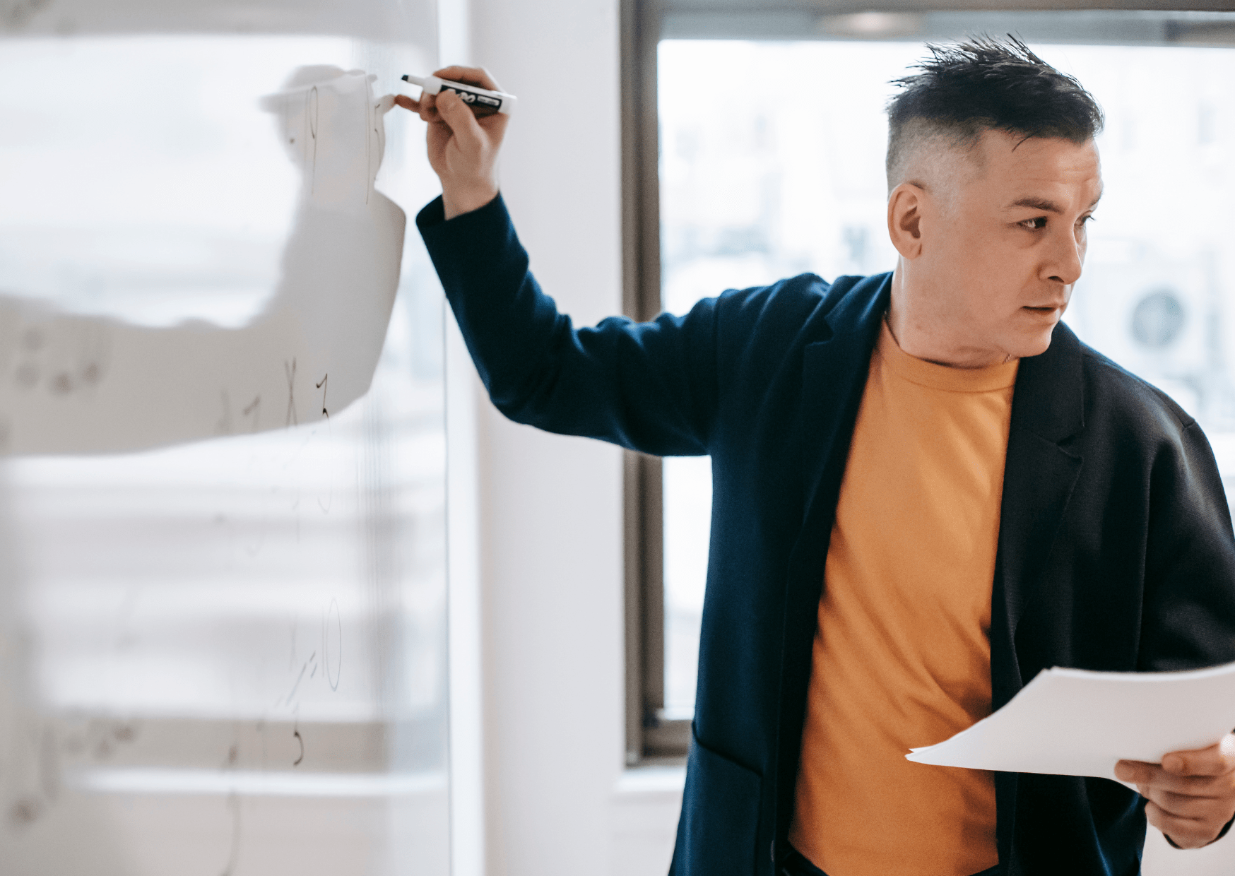 A man is writing on a whiteboard whilst looking at a recent inspection report. He is also talking to a group which can't be seen.