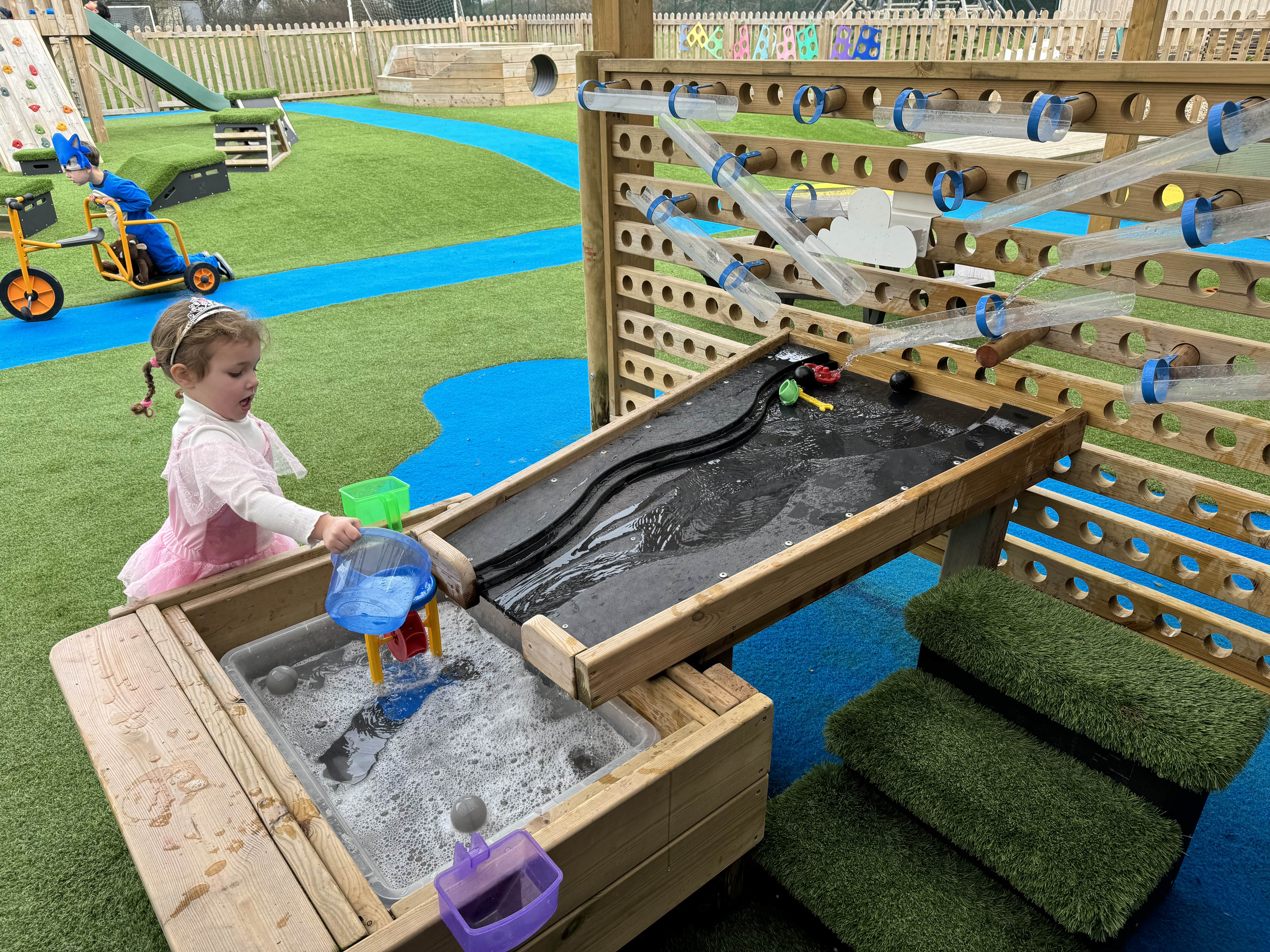 A little girl is pouring water into a water toy, which is a part of the Water Play package. The water wall and water damming table can be seen connecting to the sand pit that the little girl is interacting with. A set of Artificial Grass steps can be seen next to the equipment.