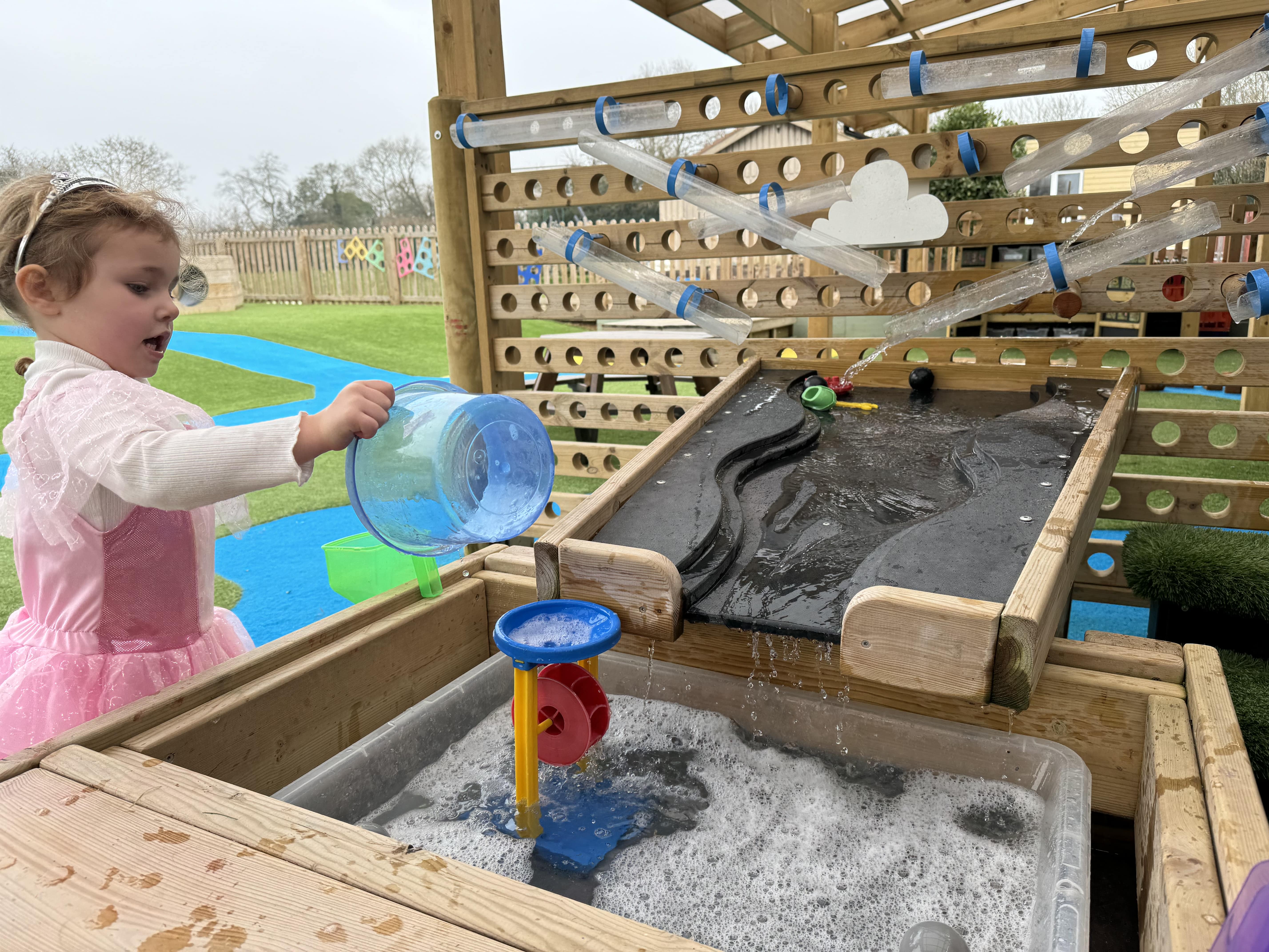 A close up of a little girl pouring water into a water play toy, which is linked to the water play package.