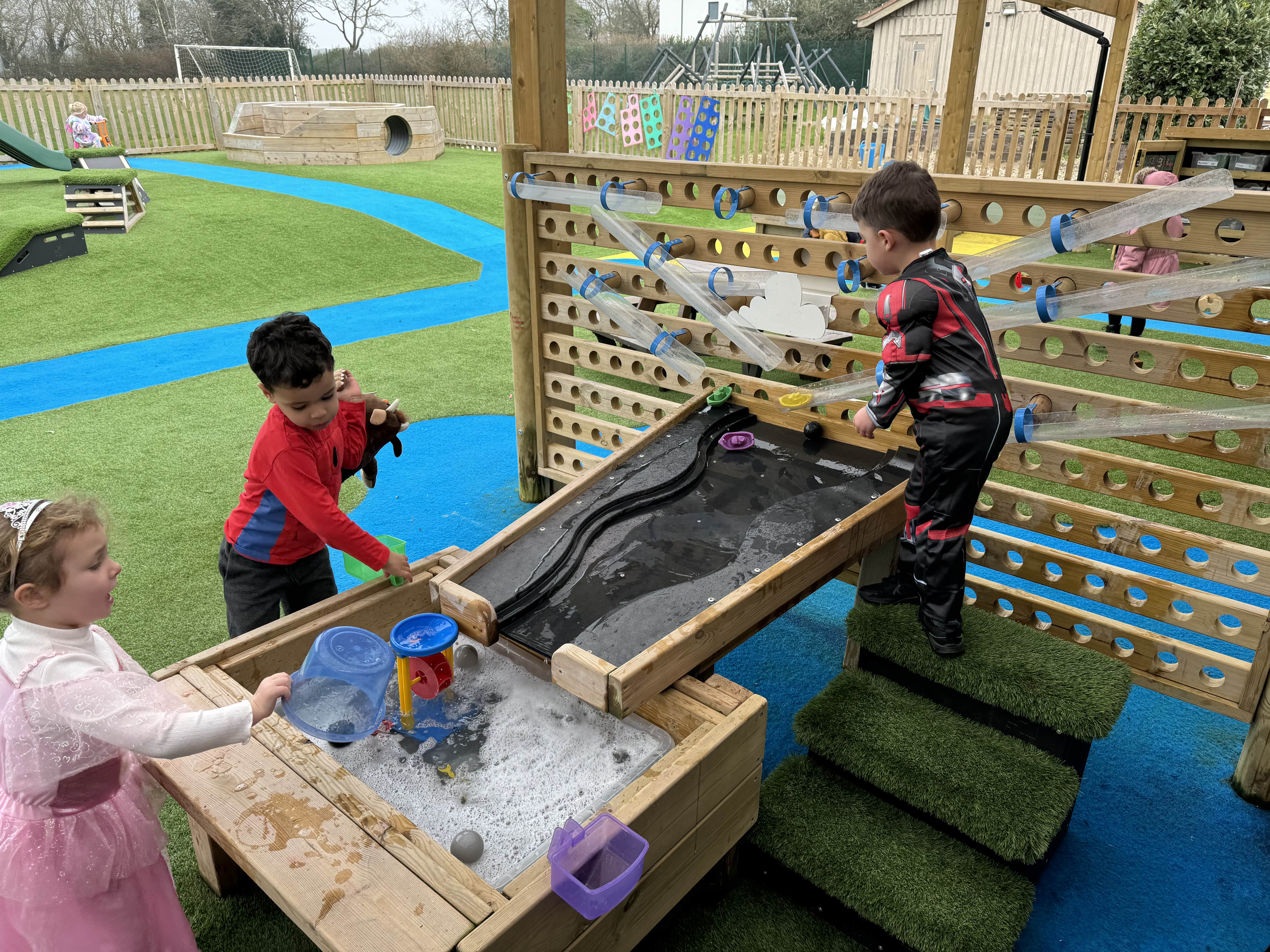 A group of children are playing with the water wall by pouring water down a pipe. The pipe end then points at a water dam table, which causes the water to poor down the table and into the sand box. 