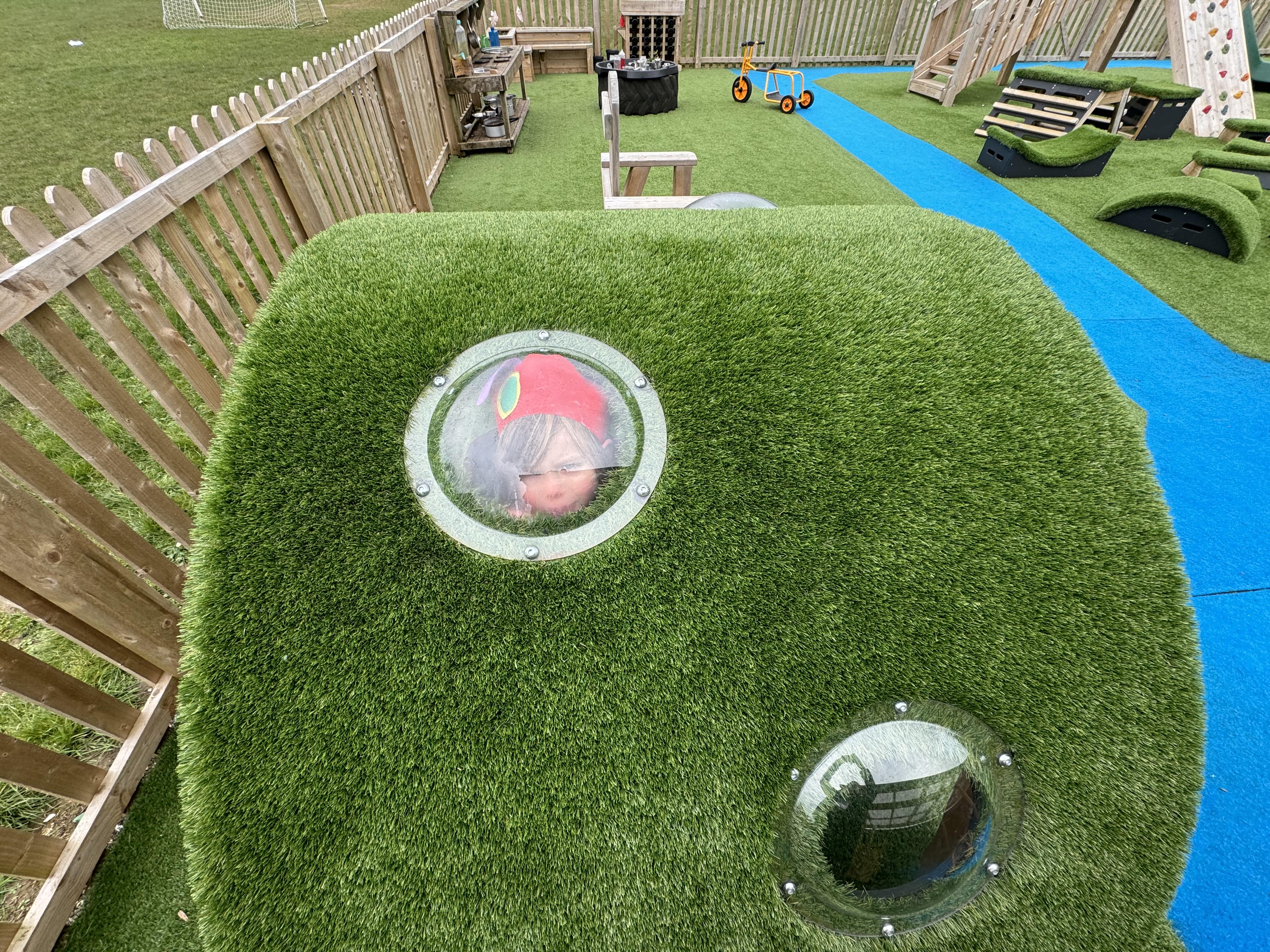 A child is looking through a small window built into the side of the Hill Den. The child is smiling whilst looking towards the camera.