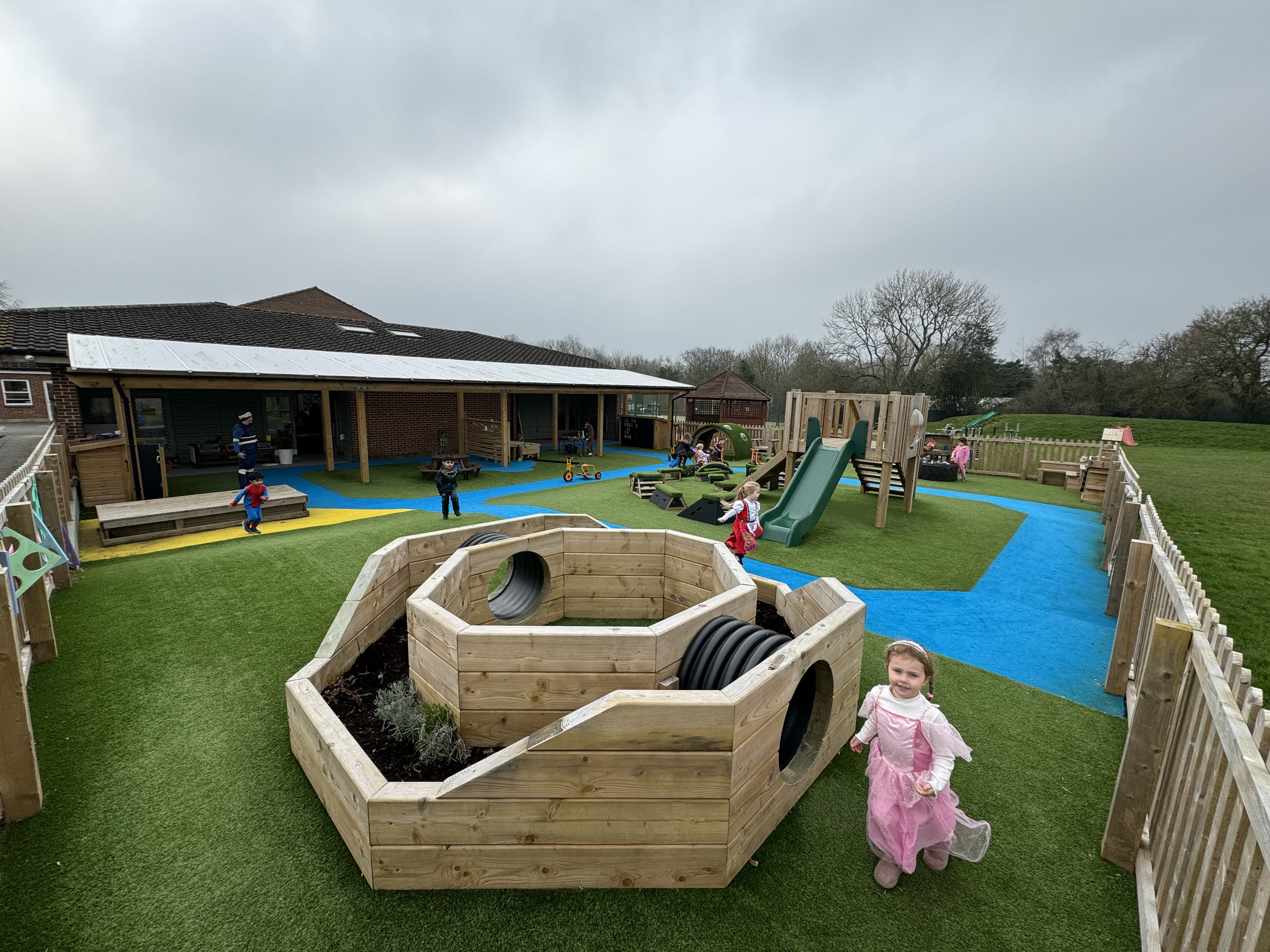 The immersive planter can be seen in the foreground, with the modular Play Tower and other pieces of play equipment being seen in the background. All of these pieces of play equipment have been installed on the playground.