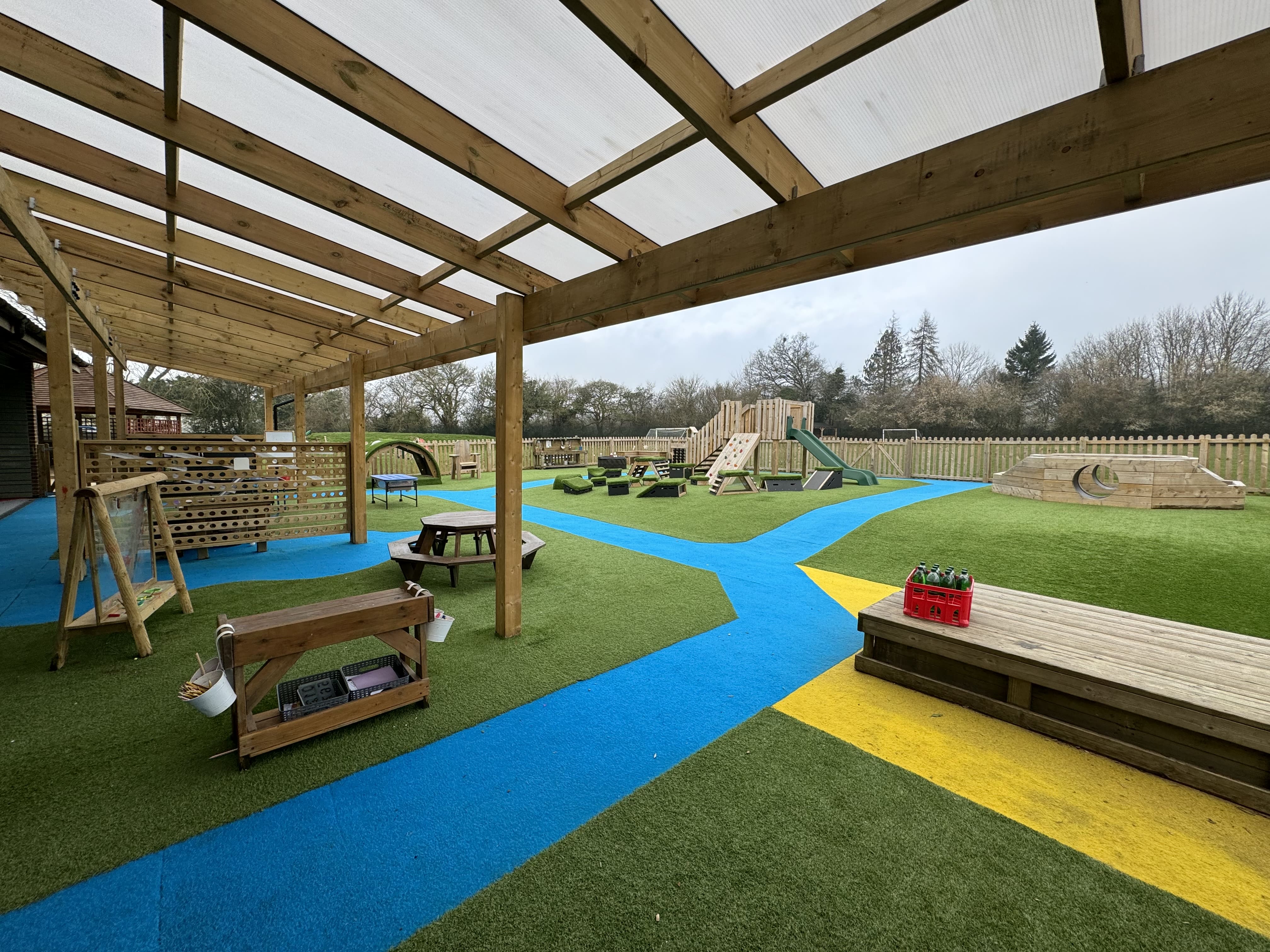 An establishing shot of the school playground with the school canopy over hanging the camera. Different pieces of play equipment can be seen around the play area.