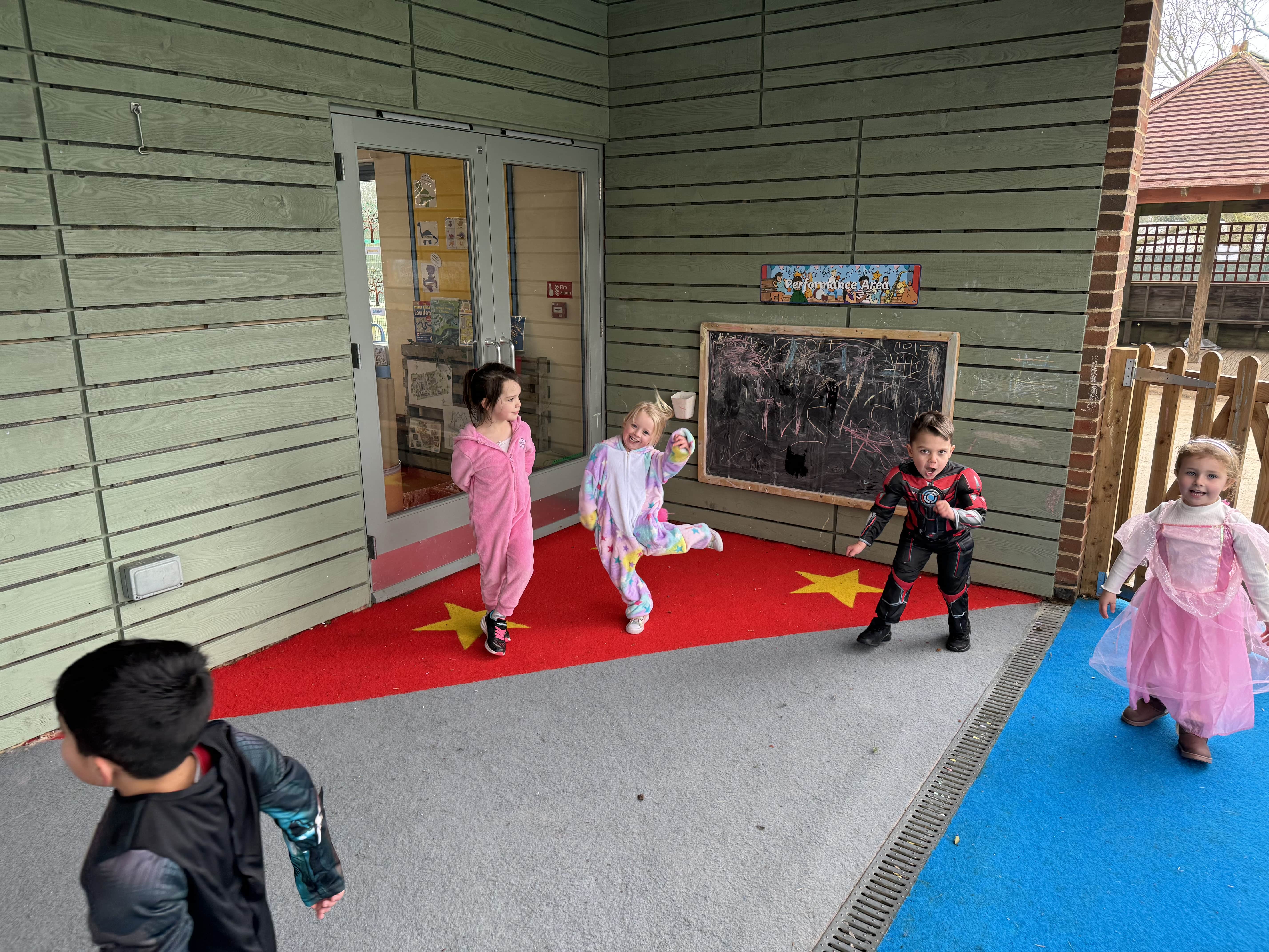 A group of children are all dancing and playing in an outdoor area with artificial grass surfacing. The grass is Red and Grey with yellow stars. A chalkboard can be seen built on the wall of a building.