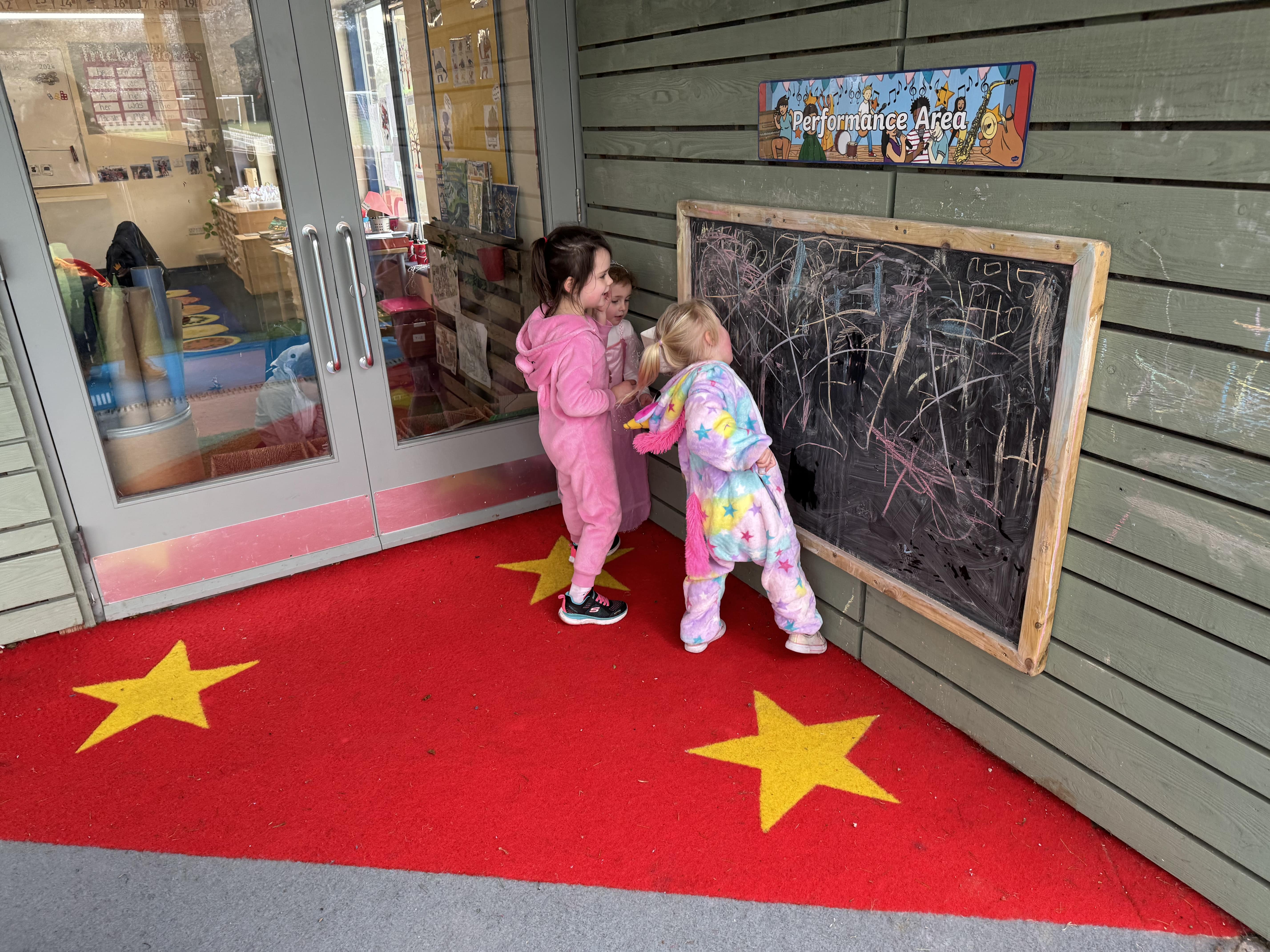 3 Children are stood around a chalkboard and are writing a variety of words and shapes on the board.