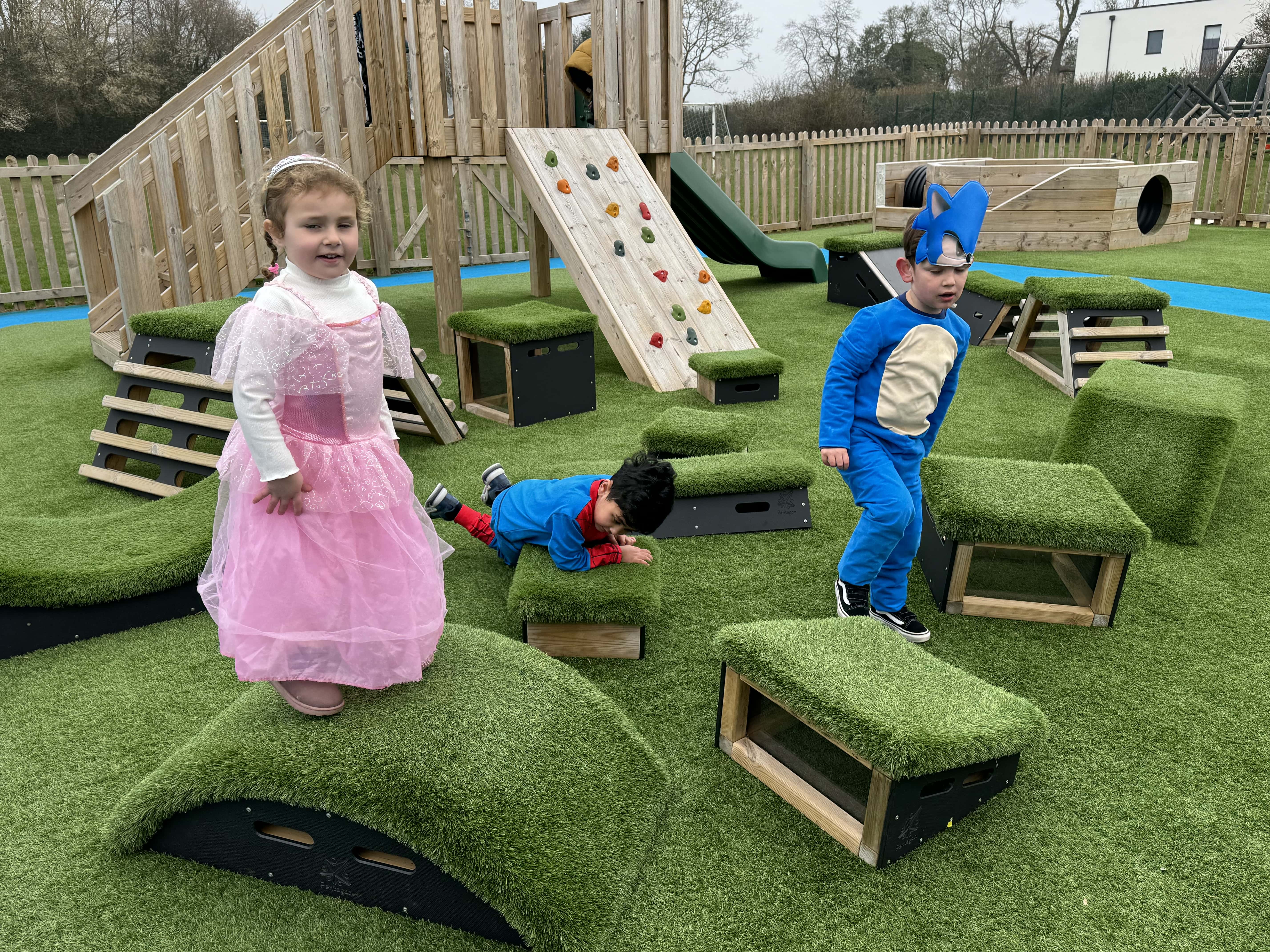 A group of children are playing on a variety of wooden blocks with artificial grass placed on top of them. A modular Play Tower can be seen in the background.