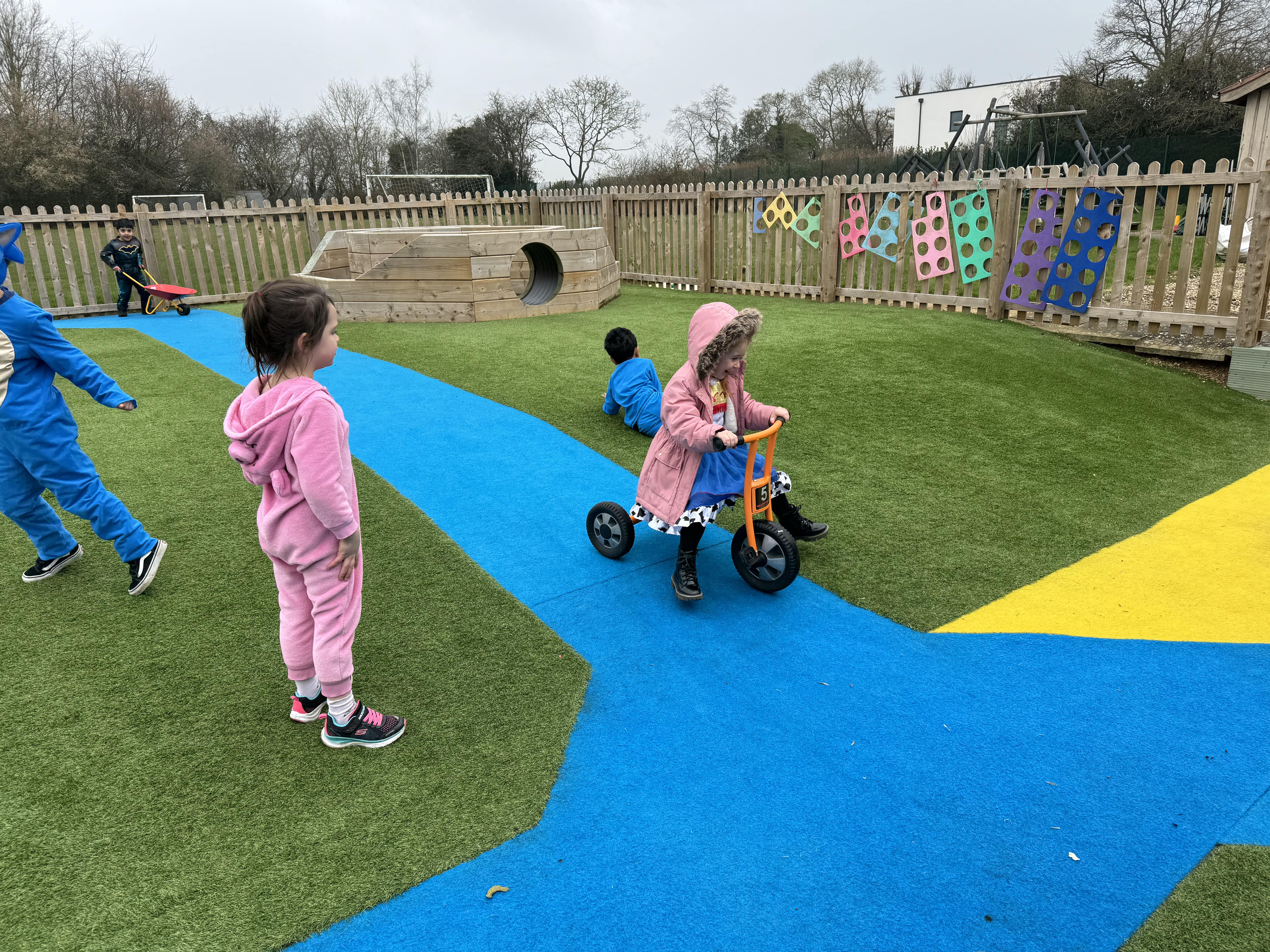 A group of children are playing on the artificial grass surfacing. A child is on a tricycle and is cycling along a blue road. Another child can be seen with a wheelbarrow in the background, wheeling it towards the camera.