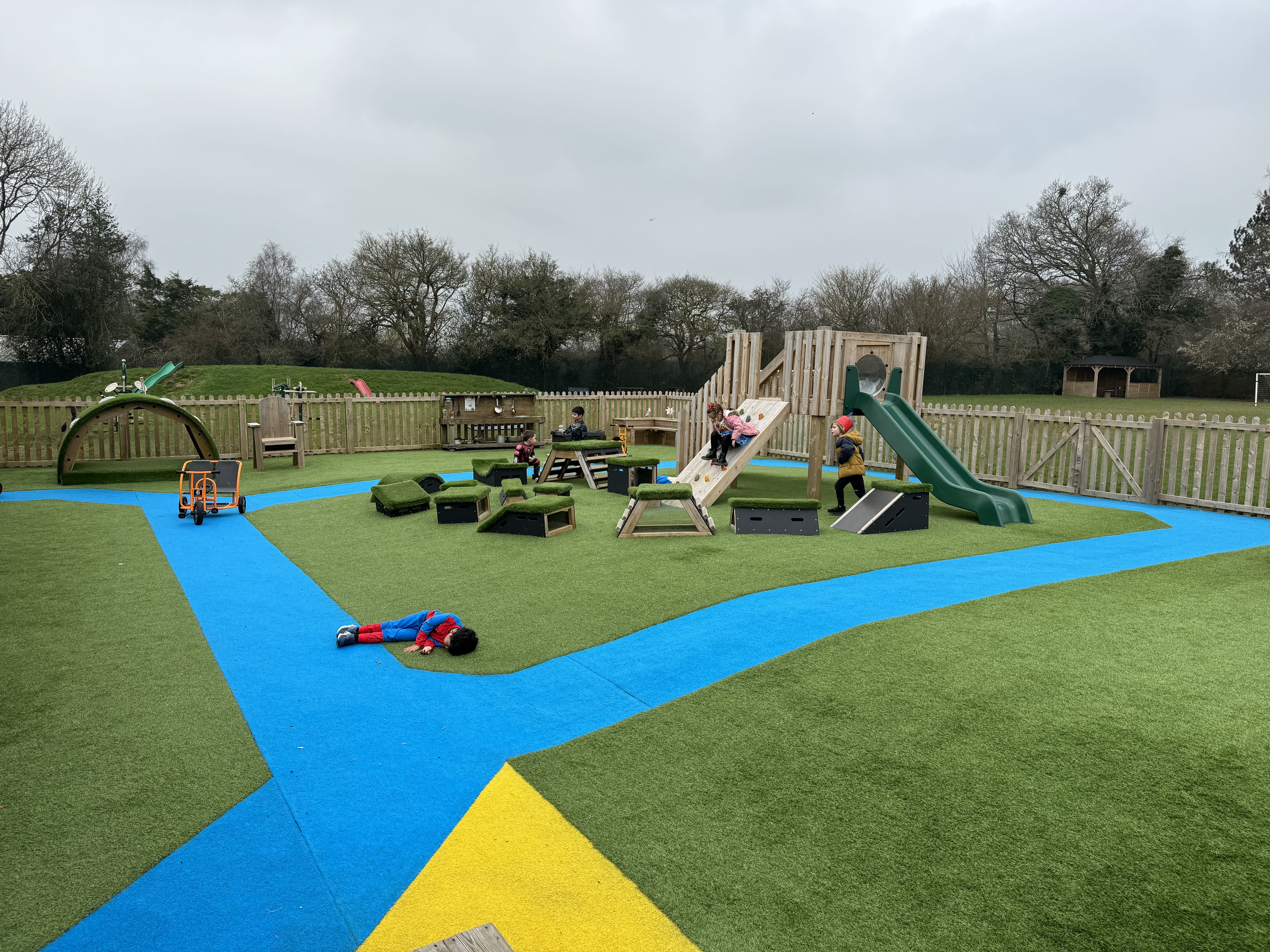 A variety of play equipment can be seen placed on the artificial grass surfacing.