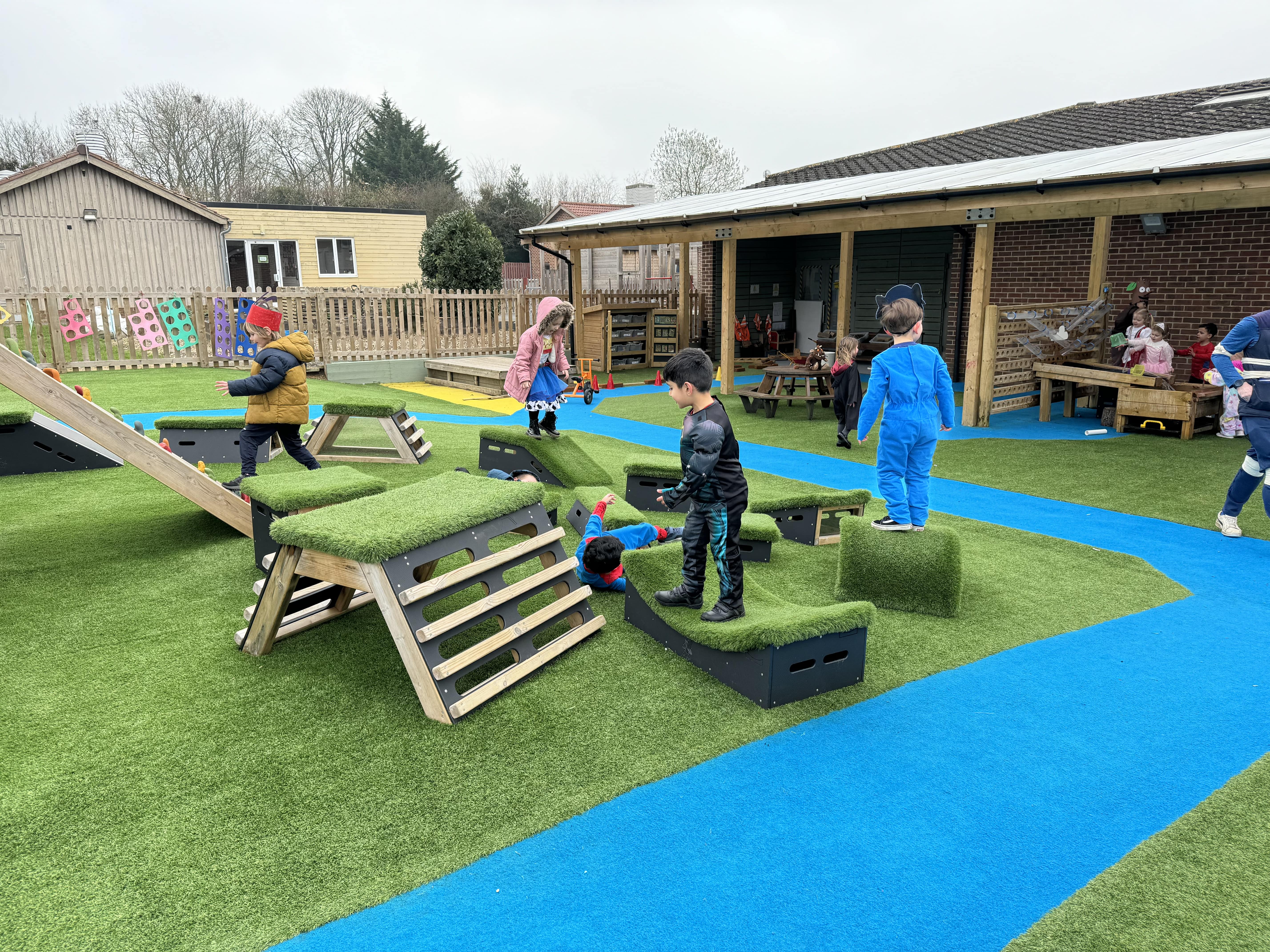 A group of kids are stood on wooden blocks which have artificial grass surfacing on the top of them. One child is jumping from a block towards a climbing ramp.